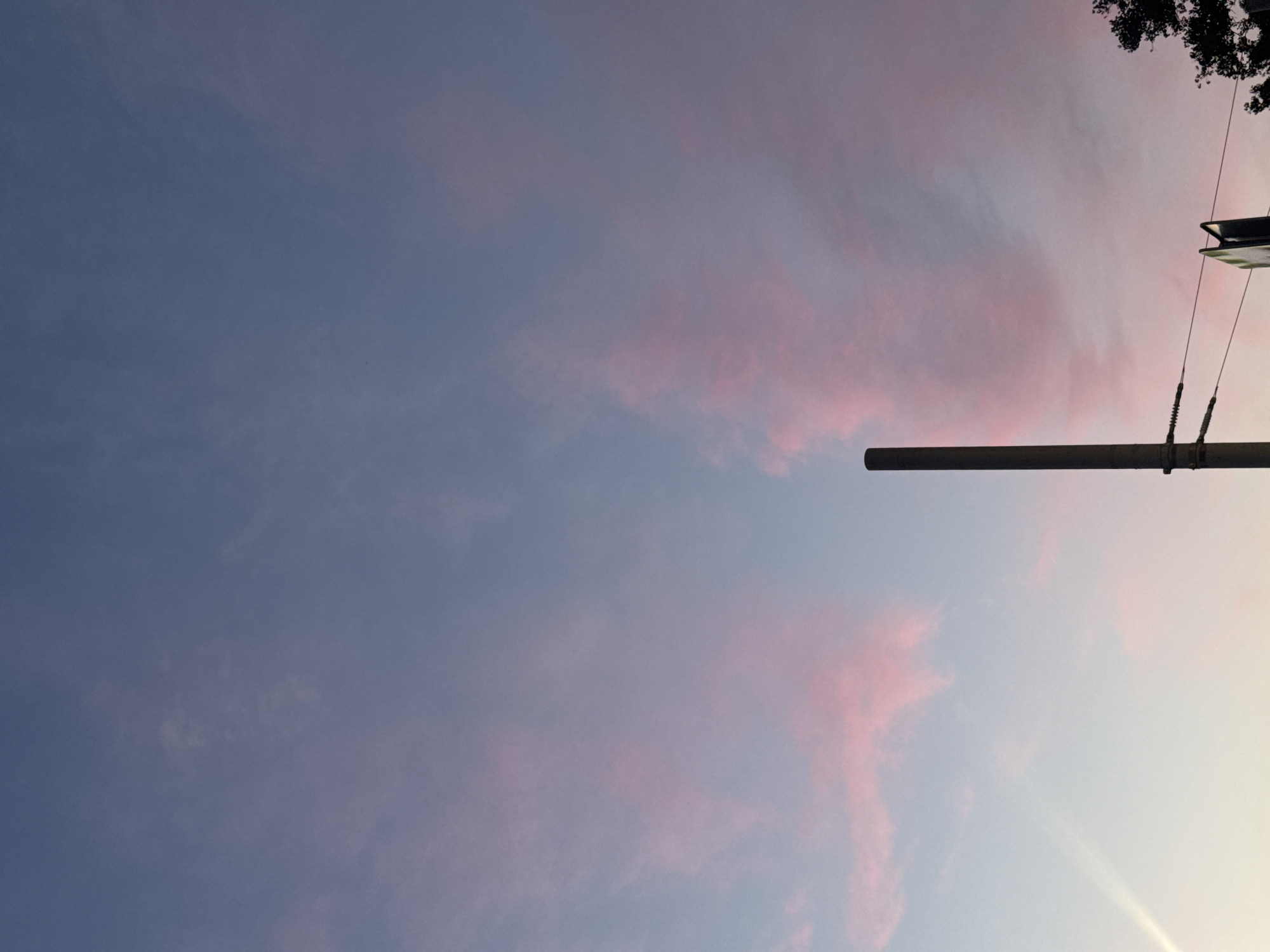 This is a photograph taken from a low angle, looking upwards towards the sky. The sky is a gradient of colors, transitioning from a deep blue on the left to a lighter, almost white hue on the right. Scattered across the sky are wisps of clouds tinged with pink, suggesting a sunset or sunrise.
In the foreground, a thick, dark horizontal pole cuts across the frame from the right side, extending towards the left. This pole appears to be part of some infrastructure, possibly a street light or a utility pole. It is supported by two coiled springs, indicating it might be a movable or flexible part of a structure. Above and slightly to the right of the pole, a black, angular object is visible, possibly the fixture of a street light or a similar apparatus. At the very top right corner, the dark leaves of a tree branch are visible, silhouetted against the sky.
The lighting suggests it is either early morning or late evening, with the sun low on the horizon, casting a soft glow and creating the pink hues in the clouds. The overall mood is calm and serene, with the vast expanse of the sky dominating the image. There is no visible text in the image. The location context is Khimki, Russia.