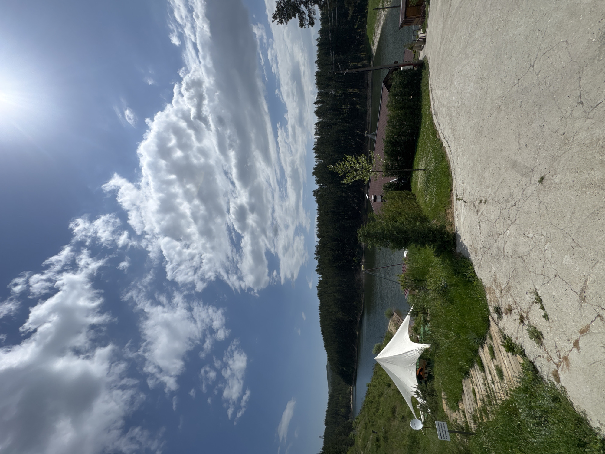 The image shows a landscape view on a sunny day. A body of water stretches between a dense forest on one side and a concrete path and grassy area on the other. A white canopy tent is erected on the grassy area. The sky is blue with scattered white clouds. It is possible this scene may be Alino, Bulgaria.
