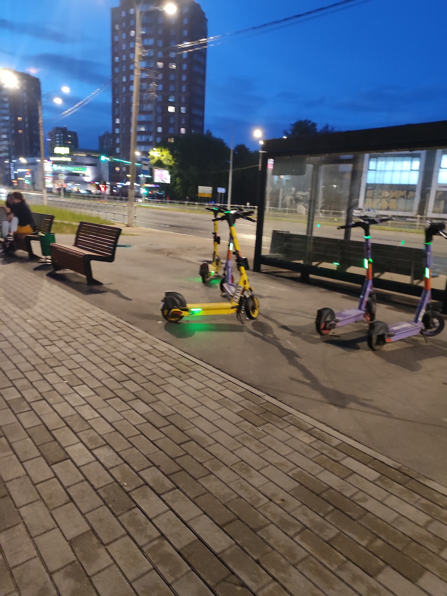 At dusk, the scene captures a city sidewalk and road. In the foreground, a paved walkway with a herringbone brick pattern runs diagonally across the frame. Adjacent to the walkway is a street with a light pole and traffic lights visible in the distance. Three electric scooters are parked on the sidewalk: a yellow one in the foreground with green lights, and two purple ones behind it, also with green lights. Behind the scooters, a bus stop shelter with glass panels and a dark roof is visible. Two benches are placed on the sidewalk near the bus stop. Further back, a multi-lane road is present with passing cars and buildings lining the street, including a tall, illuminated skyscraper. The sky is a deep blue with some clouds, indicating twilight.