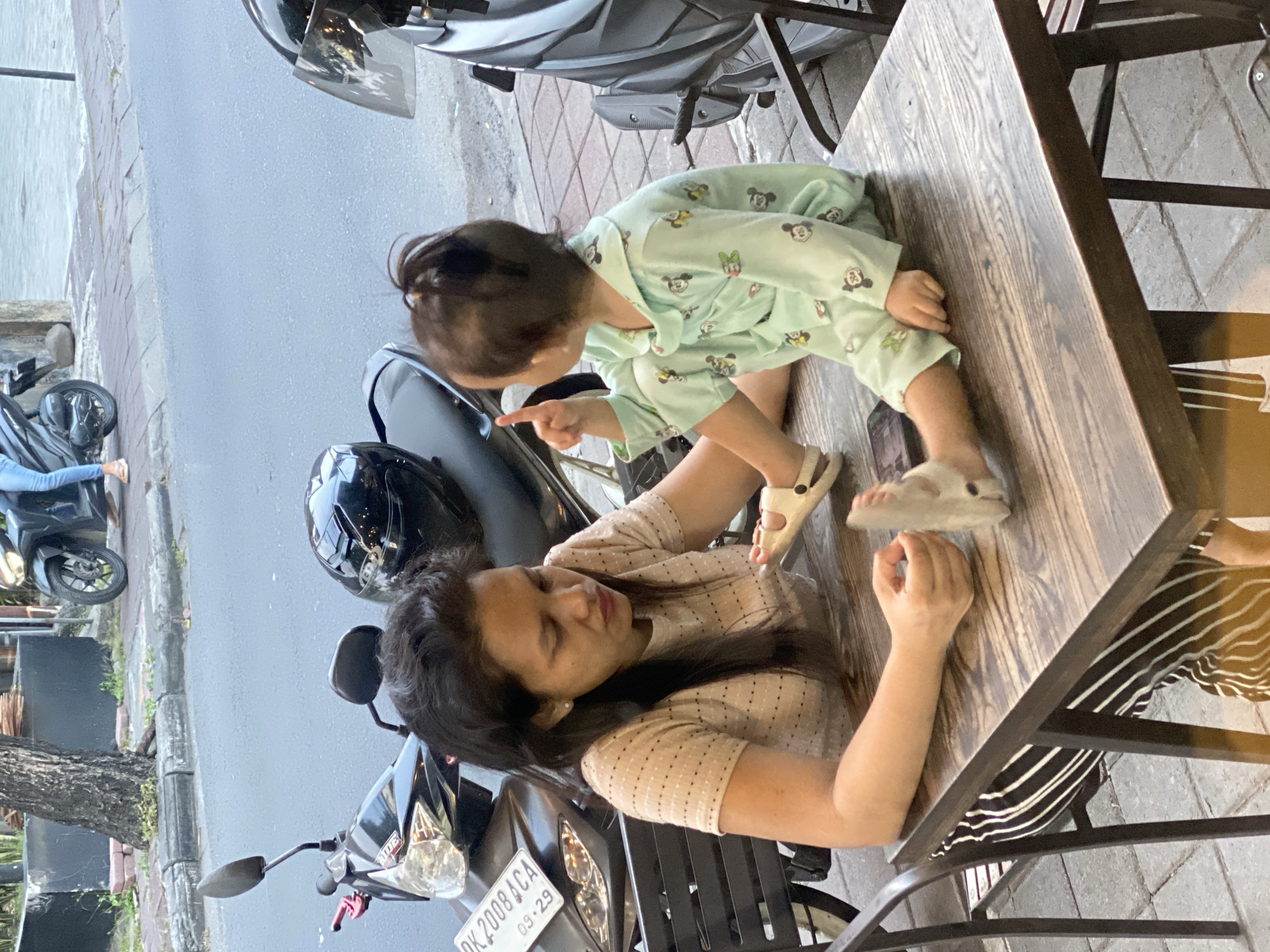 A woman and a child are seated at a wooden table outside a building in North Kuta, Indonesia. The woman, with dark hair, is wearing a light-colored, short-sleeved shirt with small dots and striped pants. The child, also with dark hair pulled into a small ponytail, is wearing a light green pajama set with a cartoon print. The child is pointing upwards with their right index finger and looking towards the left. A black helmet is resting on the seat of a motorcycle parked behind the woman. The table is made of wood with a visible grain pattern. A smartphone is partially visible on the table near the child. The ground is paved with square tiles. In the background, a motorcycle is parked, and a portion of a building is visible. The overall scene appears to be a casual outdoor setting.