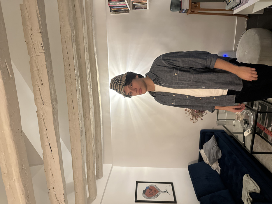 A person stands in a room featuring exposed white-painted beams on the ceiling. The individual wears a checkered hat, a white t-shirt, and a grey collared shirt. Behind them, a blue couch, a glass table, and a white ottoman are visible. A framed picture hangs on the wall, and a bookshelf is situated in the upper background. The room is brightly lit.