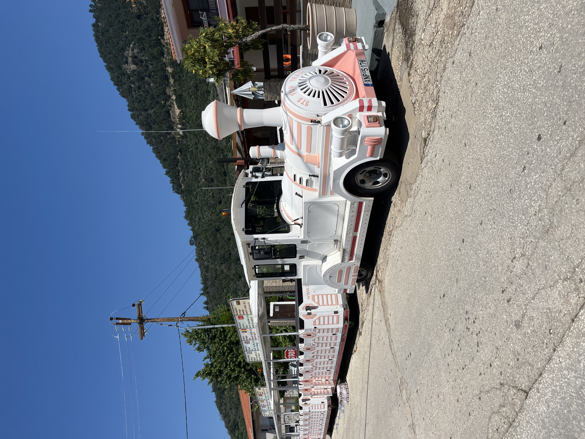 An outdoor, eye-level, full shot captures a white and pink tourist train parked on a paved street. The train consists of a locomotive and several passenger cars, all featuring a predominantly white exterior with pink accents. The locomotive is at the forefront of the image, displaying the words "STS FUN TRAIN" on its front. A license plate on the rear of the locomotive reads "XNP 5178". The passenger cars are connected in a line behind the locomotive, each with open sides and a white roof. Signs are mounted above the passenger cars, displaying information in multiple languages. A red "STOP" sign is visible on the side of one of the passenger cars. The street is made of asphalt with some cracks and dark spots. In the background, a lush green hillside rises, covered with trees. Buildings with terracotta roofs and white walls are visible on the right side of the image, along with a potted tree. A utility pole with wires stands on the left side of the frame. The sky is clear and blue.
