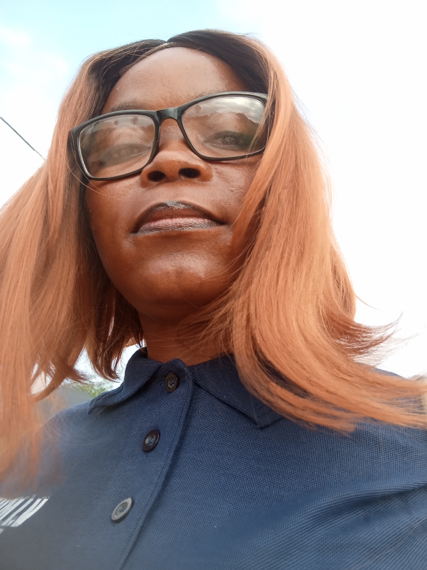 A close-up, low-angle selfie shows a Black woman with medium-toned skin and reddish-brown hair wearing black-rimmed glasses and a dark blue collared shirt. She has dark eyeliner and lip gloss on. Her hair is straight and falls to her shoulders. The background is a bright, overcast sky with some faint clouds. The scene appears to be outdoors, possibly in Kason, Nigeria.