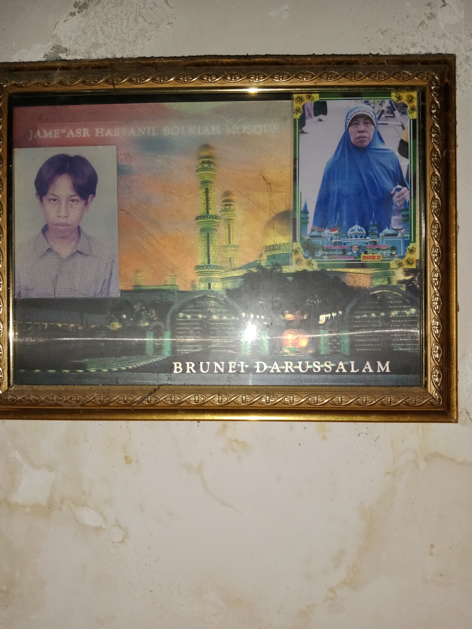 The image is a framed photograph featuring two portraits and a mosque in the background, likely taken in Ringinrejo, Indonesia. The left side of the frame displays a young man wearing a checkered shirt, positioned against a backdrop of a mosque with tall, green minarets and golden domes. Text above this portrait reads "JAME"ASR HASSANIL BOLKIAH MOSQUE". The right side of the frame shows an older woman wearing a blue hijab, also against a similar mosque backdrop. Below the mosque scene, the words "BRUNEI DARUSSALAM" are visible, suggesting the mosque is located there. The frame itself is ornate and golden. The photograph appears aged and possibly damaged, with some discoloration and smudges on the surface. The background wall is textured and appears to be painted a light color, with some staining and peeling.