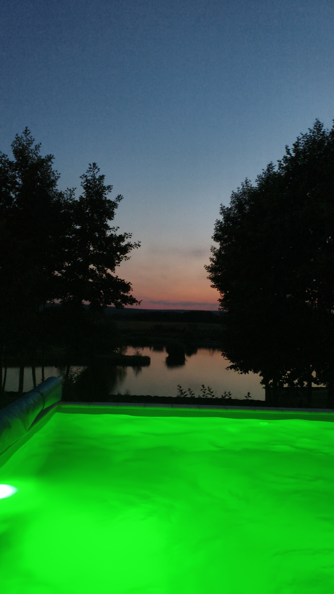 An outdoor scene at dusk features a foreground filled with a bright green, illuminated swimming pool. The pool's surface shows gentle ripples, and a small, bright light source is visible on the left edge. Beyond the pool, silhouetted trees frame a tranquil lake or pond. The water reflects the dim sky and surrounding landscape. In the distance, a faint sunset displays hues of orange and pink against a darkening blue sky, hinting at the rural outskirts of Bezliudivka, Ukraine. The overall atmosphere is serene and twilight-like.