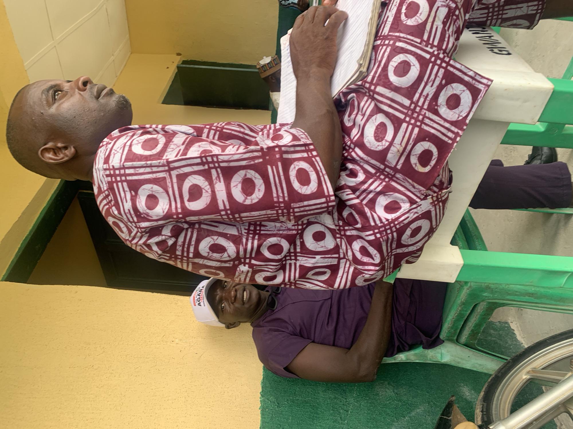 The image shows two men in Tabu, Nigeria. One man, dressed in a maroon and white patterned shirt, sits on a green plastic chair with a white top. He holds a notepad. He is looking up. Below him, another man in a purple shirt and a white "AGARA" cap sits on a green plastic chair with a white top and looks up at the man above. The scene takes place outside a yellow building with green trim. A bicycle wheel is visible to the right.
