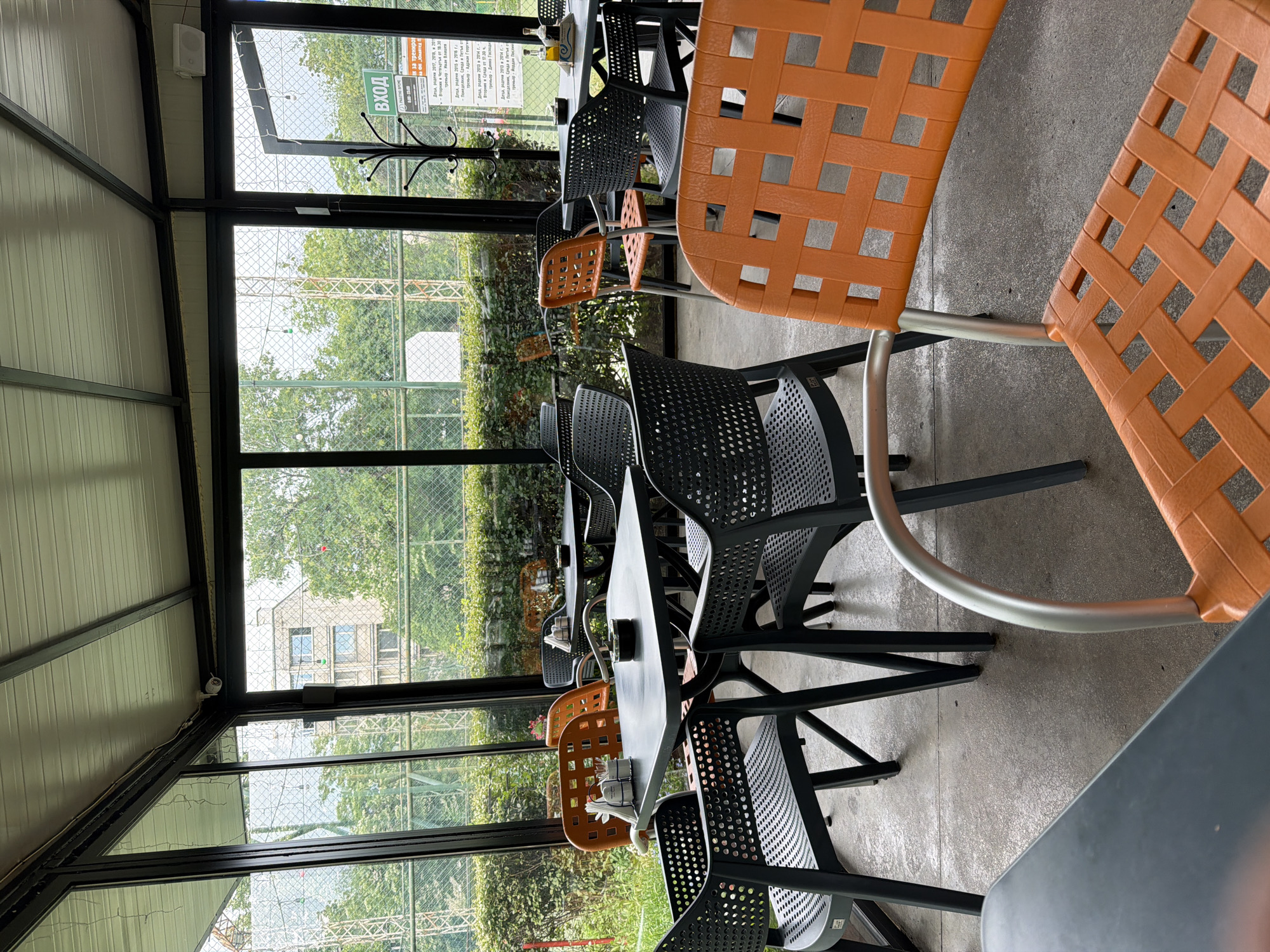This media file captures an empty indoor dining or cafe area, part of what appears to be a sports facility in Plovdiv, Bulgaria. The space is bright, illuminated by natural light filtering through large, floor-to-ceiling glass windows that form one wall of the room. The ceiling and inner walls are covered with white, horizontally paneled material.
The room is furnished with multiple dark gray/black tables and chairs. Some chairs are made of black perforated plastic, while others are a vibrant orange with a woven lattice pattern on their backs and seats, adding a splash of color to the otherwise monochrome furniture. On some tables, white napkins and small containers are visible, suggesting the space is prepared for guests. A black metal coat rack stands near the window, and a white speaker and a security camera are mounted on the ceiling or wall. The floor is made of concrete.
Beyond the windows, a lush, green outdoor environment is visible, dominated by trees and dense foliage. A chain-link fence runs along the perimeter, indicating a sports court or field just outside. In the background, through the greenery, a light-colored building with windows can be vaguely seen. No people are present, lending a quiet, expectant atmosphere to the scene.
Visible text on a sign outside the window, in Bulgarian, reads "ВХОД" (Entrance). Further text on what appears to be a schedule or information board details "тренировки" (training sessions) for children, specifying age groups (e.g., "Деца, родени 2017, 2018, и 2011" - Children, born 2017, 2018, and 2011), days ("Вторник и Четвъртък" - Tuesday and Thursday), times (e.g., "от 18:30" - from 18:30), and coaches' names (e.g., "Адриан Георги", "Динко Господи", "Йордан"). This confirms the setting is likely a lounge or cafe associated with a children's sports club or training center.