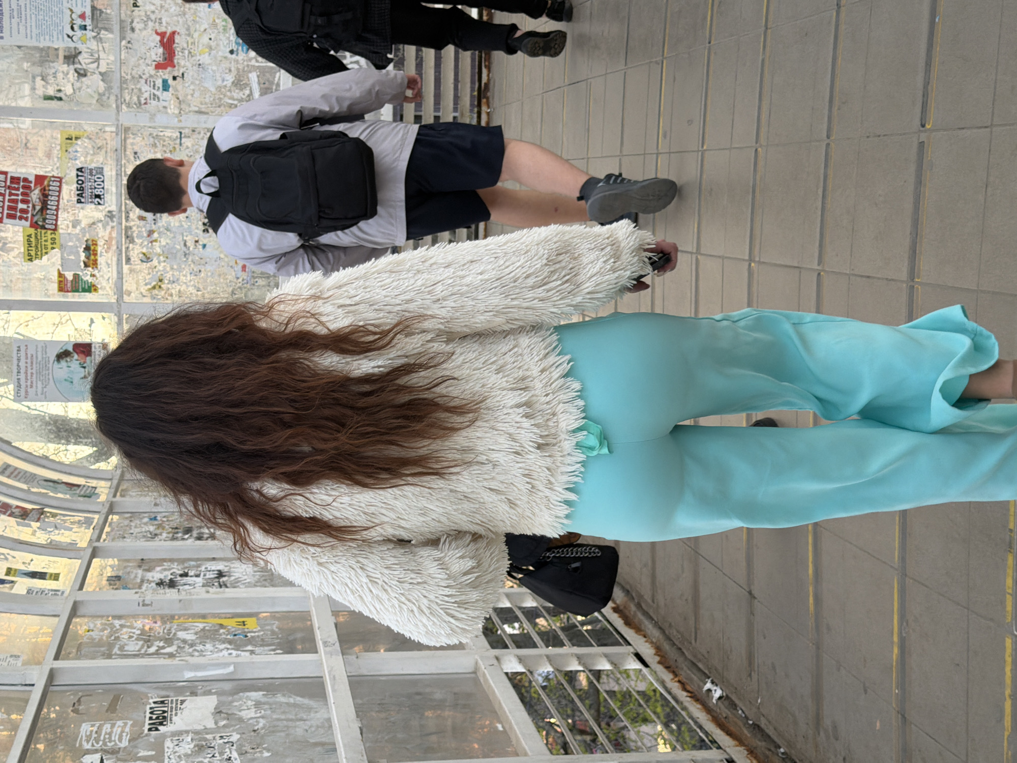 The image captures an outdoor scene on a public walkway or underpass in Beryozovy, Russia, during the daytime. The perspective is from behind a person with long, wavy brown hair, who is the main subject. This individual wears a cream or off-white fluffy jacket and light blue, possibly teal, wide-leg pants, and carries a small black handbag with a chain strap.
Ahead and slightly to the right, another person walks in the same direction, also seen from the back. They are dressed in a light grey long-sleeved shirt, dark shorts, and black sneakers, carrying a black backpack. Further in the background, a third person in dark attire is partially visible. The ground is a light grey tiled surface with thin yellow lines.
To the left, a structure composed of framed panels, likely glass or plastic, is heavily plastered with various paper advertisements and flyers. The visible Russian text on these posters includes "РАБОТА" (Work), "СТУДИЯ ТВОРЧЕСТВА" (Creativity Studio) advertising "Курсы кройки и шитья" (Sewing and tailoring courses), and an offer for "АРТИРА 20.000Р" (Apartment for 20,000 Rubles). Several phone numbers, such as "89094660861" and "8-964-91-77-000," are also present, along with the number "2.800 P" and the word "DATA." The scene depicts everyday pedestrian activity in a public urban environment.