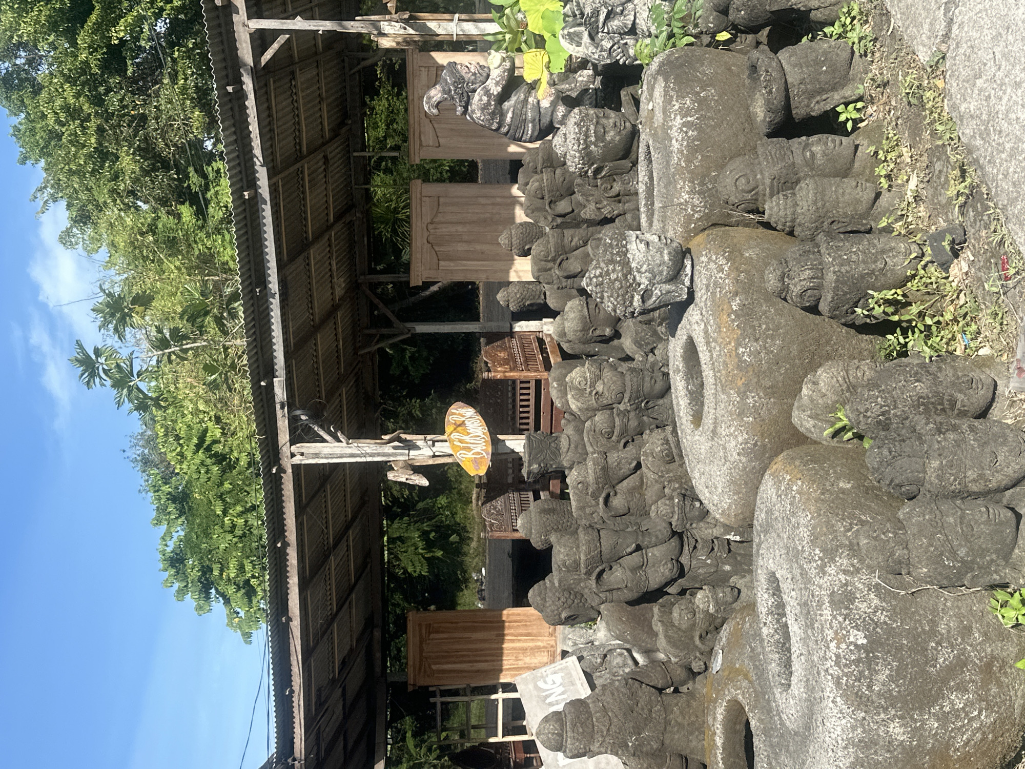 This outdoor scene features a collection of stone sculptures displayed under a weathered corrugated metal roof supported by wooden posts. In the foreground, numerous carved stone heads and torsos, many with headdresses, are arranged on the ground. Several large, circular stone containers with openings in the center are prominent among the smaller sculptures. In the background, large wooden doors are visible, along with some ornate wooden furniture. A vibrant yellow oval sign with the text "Bali Sunshine" hangs from a wooden post. A dark, figurative stone statue stands to the right of the wooden doors, partially obscured by lush green foliage. The ground is a mixture of dirt and concrete. The sky above is clear and blue. The location is identified as Denpasar, Indonesia.