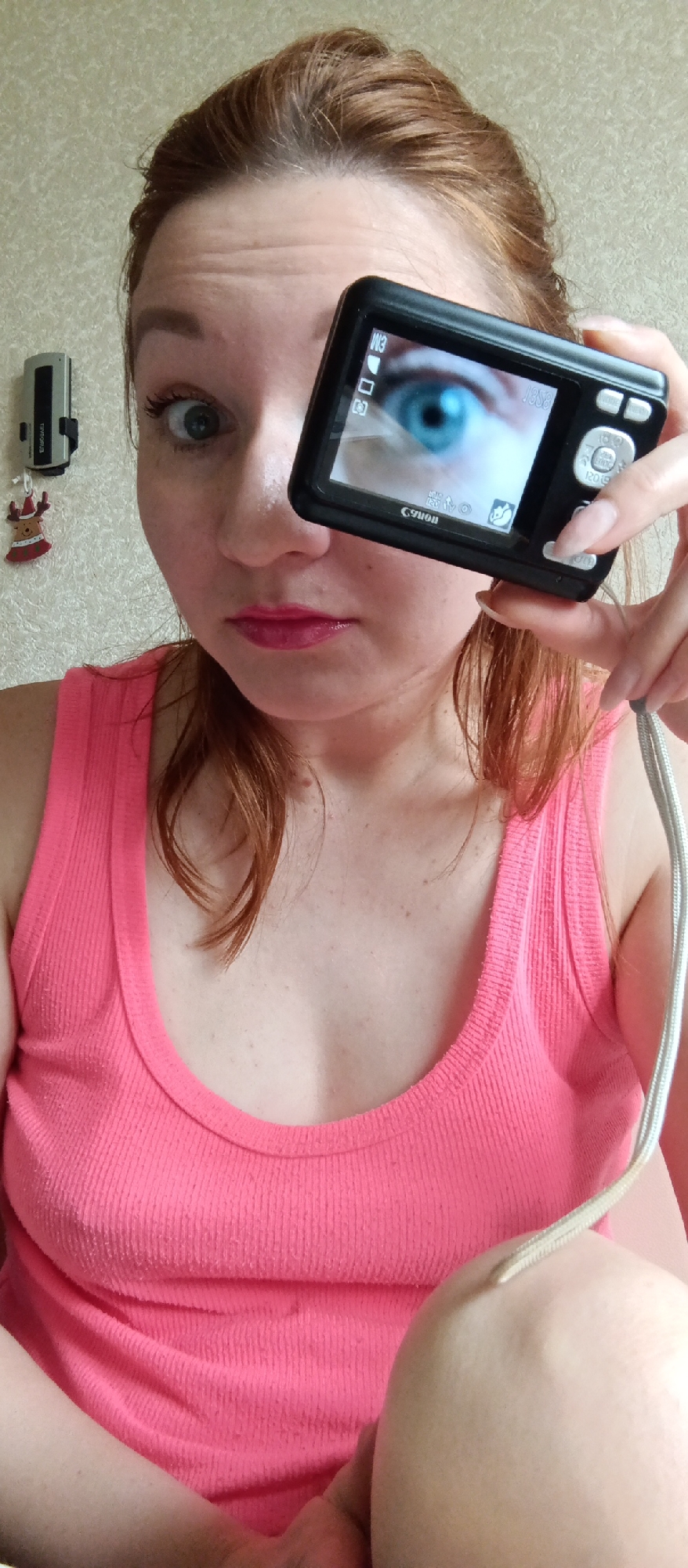 A woman with reddish-brown hair pulled back, wearing a pink tank top, is holding a black Canon digital camera in front of her face. The camera screen displays a close-up of a bright blue eye. Her left eye is visible, showing a surprised expression. The background is a wall with a small, silver-colored appliance and a Christmas ornament hanging on it. The location is Unknown, Unknown.