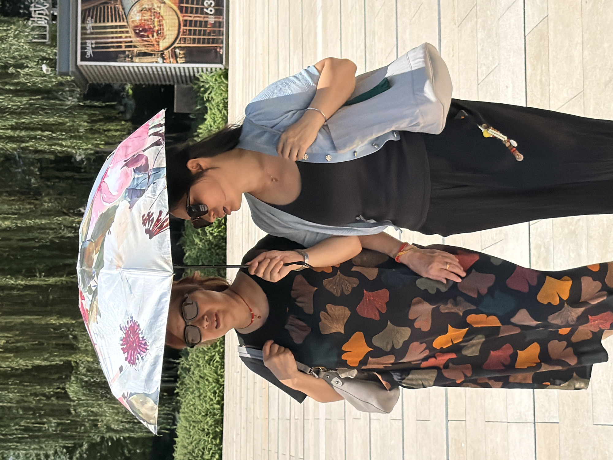 Two women are walking side-by-side under a patterned umbrella. The woman on the left wears a black dress with a ginkgo leaf print in shades of orange, red, brown, and green. She has short, brown hair, wears glasses, and has a red necklace. A gray shoulder bag hangs from her left shoulder. The woman on the right has long, dark hair and wears a light blue cardigan over a black tank top and black pants. She has sunglasses on and wears a silver bracelet. A light blue tote bag is slung over her right shoulder. Both women are holding the umbrella. They are on a paved walkway with a line of bushes behind them, and trees with long, drooping branches further in the background. To the right, a building with a glass facade and signage is visible, possibly in a commercial area. The scene appears to be outdoors on a sunny day.