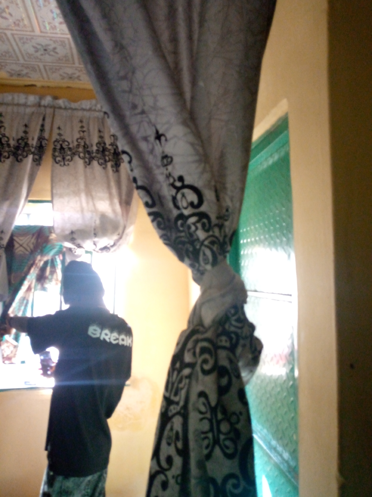 The image depicts an indoor scene, likely a room, with a person standing near a window. The person is wearing a black t-shirt with the word "BREAK" printed on the back in white letters, and patterned trousers. The person is facing away from the camera and towards the window, with their left arm extended.
In the background, there is a window with a green, textured glass pane, and sheer curtains with a black decorative pattern are visible, framing the window. A large, patterned curtain, also with black designs, hangs prominently in the foreground, partially obscuring the view. The ceiling above appears to be tiled. The walls are painted in a light, warm color. The lighting suggests it is daytime, with light coming through the window.
The specific location in the city of Jalingo, Nigeria, cannot be determined from the visual information provided in the image.