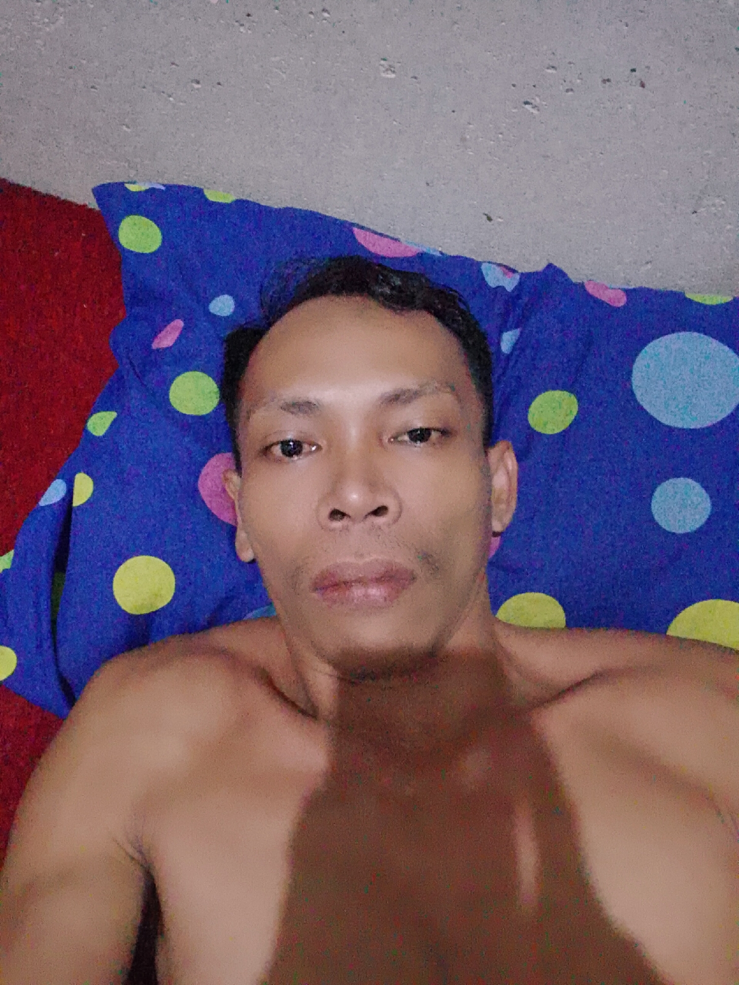 A shirtless man lies on a blue pillow with colored dots, with a red fabric to the left, and a grey wall above. The location is Bukit Ketok, Indonesia.