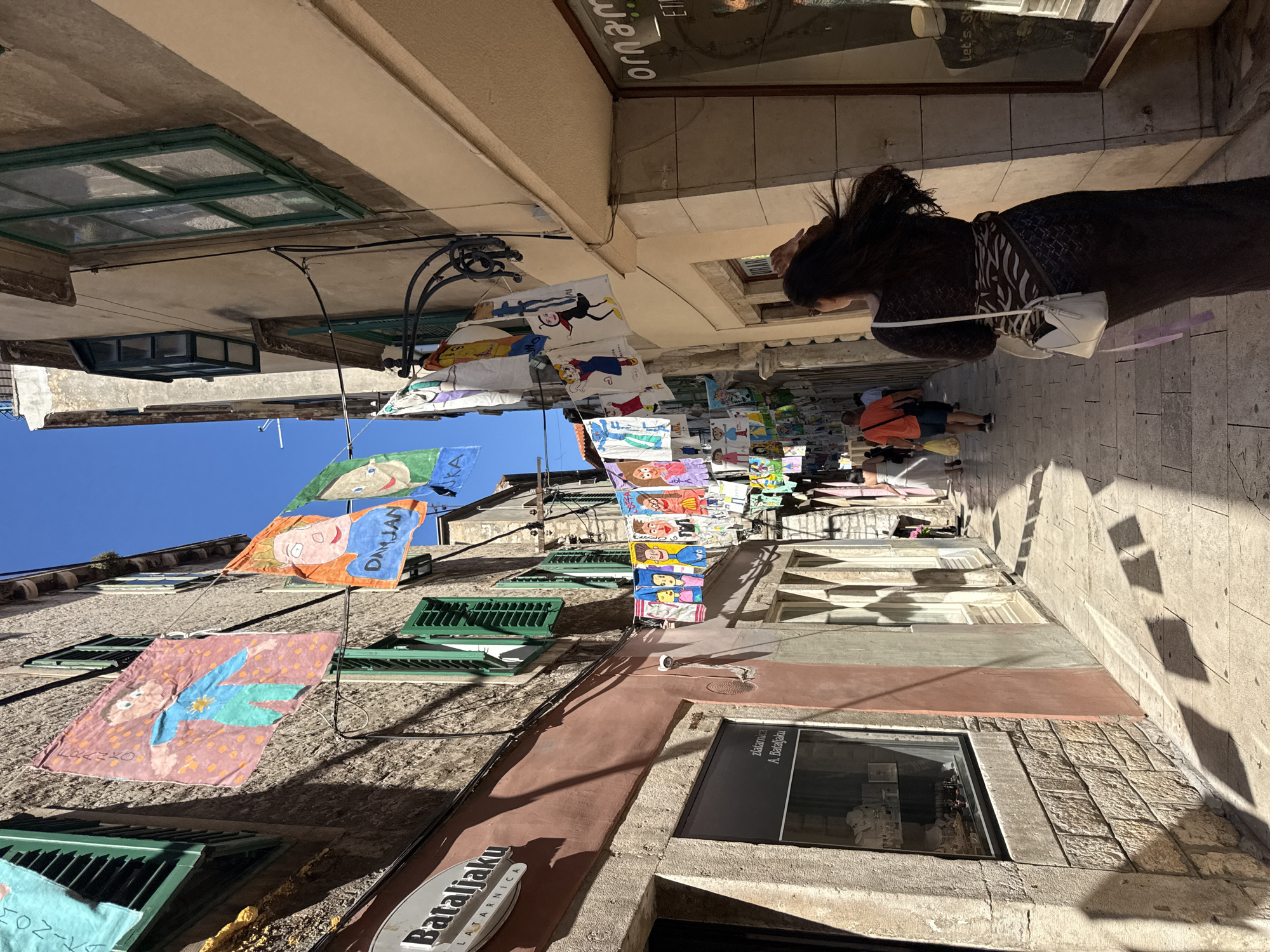 The image depicts a narrow, sun-drenched street in the historic city of Šibenik, Croatia, possibly on Mihovila Klaića. The street is lined with old stone buildings, their facades adorned with green shutters. A variety of children's drawings, presented as colorful banners, are strung across the street, creating a festive and vibrant atmosphere. Some of the drawings feature cartoonish figures, with names like "Franjo" and "Damjan" visible on some.
In the foreground, a woman with long, dark hair, wearing a dark, patterned dress and a white crossbody bag, walks away from the camera, heading down the street. Her hair is windswept. To the left of the street, a shop window displays jewelry, with a sign that reads "Batalijaku LATARNIJA". Further down the street, several people are visible, including a man in an orange shirt carrying a yellow bag, and other figures walking in both directions. Shadows of the buildings and possibly some of the banners are cast across the cobblestone pavement. On the right side of the street, another shop window, with the partially visible word "ornam..." possibly indicating an ornament shop, reflects the street scene. The sky is a clear, bright blue.