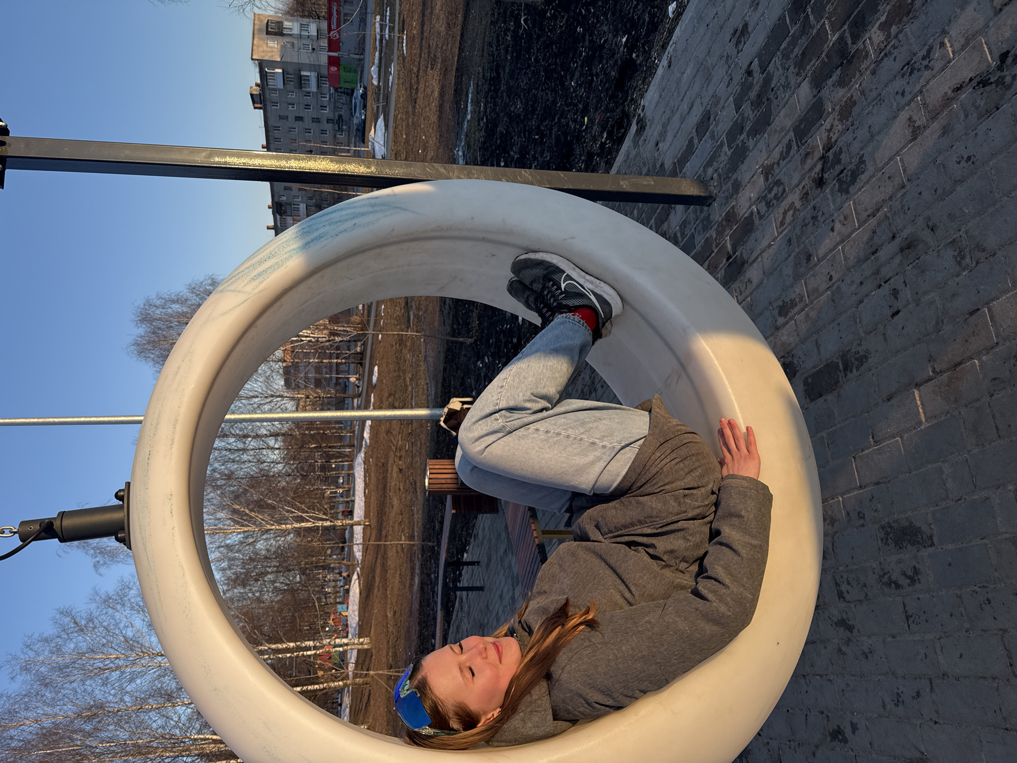 A girl lies in a white, circular swing set structure in what appears to be a park or recreation area. She is wearing a gray hooded jacket, light blue jeans, and black sneakers with red socks. Her head is to the bottom of the frame, and her feet are towards the top. Her eyes are closed and she seems to be resting or relaxing. The swing is attached to a black metal frame.
In the background, there are trees without leaves and patches of snow on the ground, suggesting it is late winter or early spring. There is a multi-story building visible in the distance. A paved area of dark bricks is to the right of the swing.
While I cannot specifically identify the exact location as Berezniki, Russia, the architecture and environment are consistent with that region.