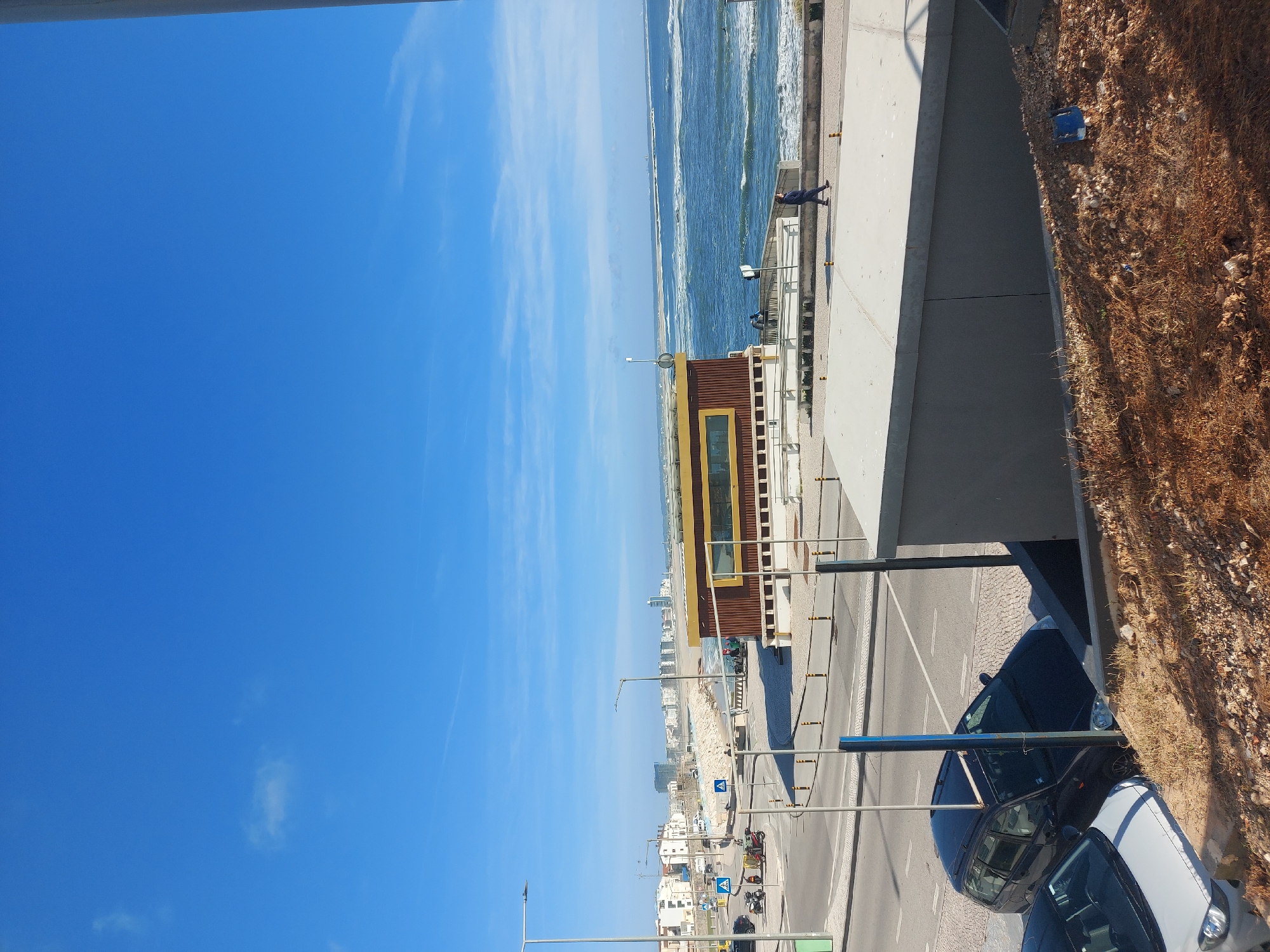 The image depicts a coastal scene in Figueira da Foz, Portugal. A clear blue sky with scattered clouds dominates the upper portion of the frame. Below, the Atlantic Ocean stretches along the horizon, displaying visible wave patterns. A concrete boardwalk runs parallel to the beach. A single person is visible on the boardwalk. Adjacent to the boardwalk is a multi-story structure with yellow trim, possibly a lifeguard station or recreational facility. A paved road runs alongside this structure. Parked cars are visible along the road. Buildings are visible in the background, along the beach.