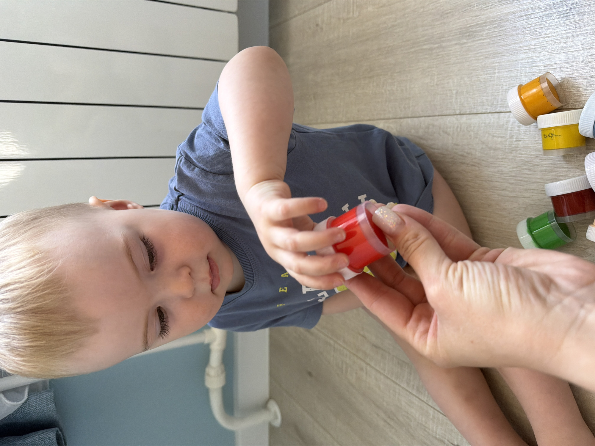 A young child is sitting on a wooden floor, reaching for a small container of red paint being held by an adult's hand. Several other containers of paint in various colors are scattered nearby. The scene is indoors, with a white wall and pipe visible in the background. There is no indication of this image being taken specifically in Votkinsk, Russia.