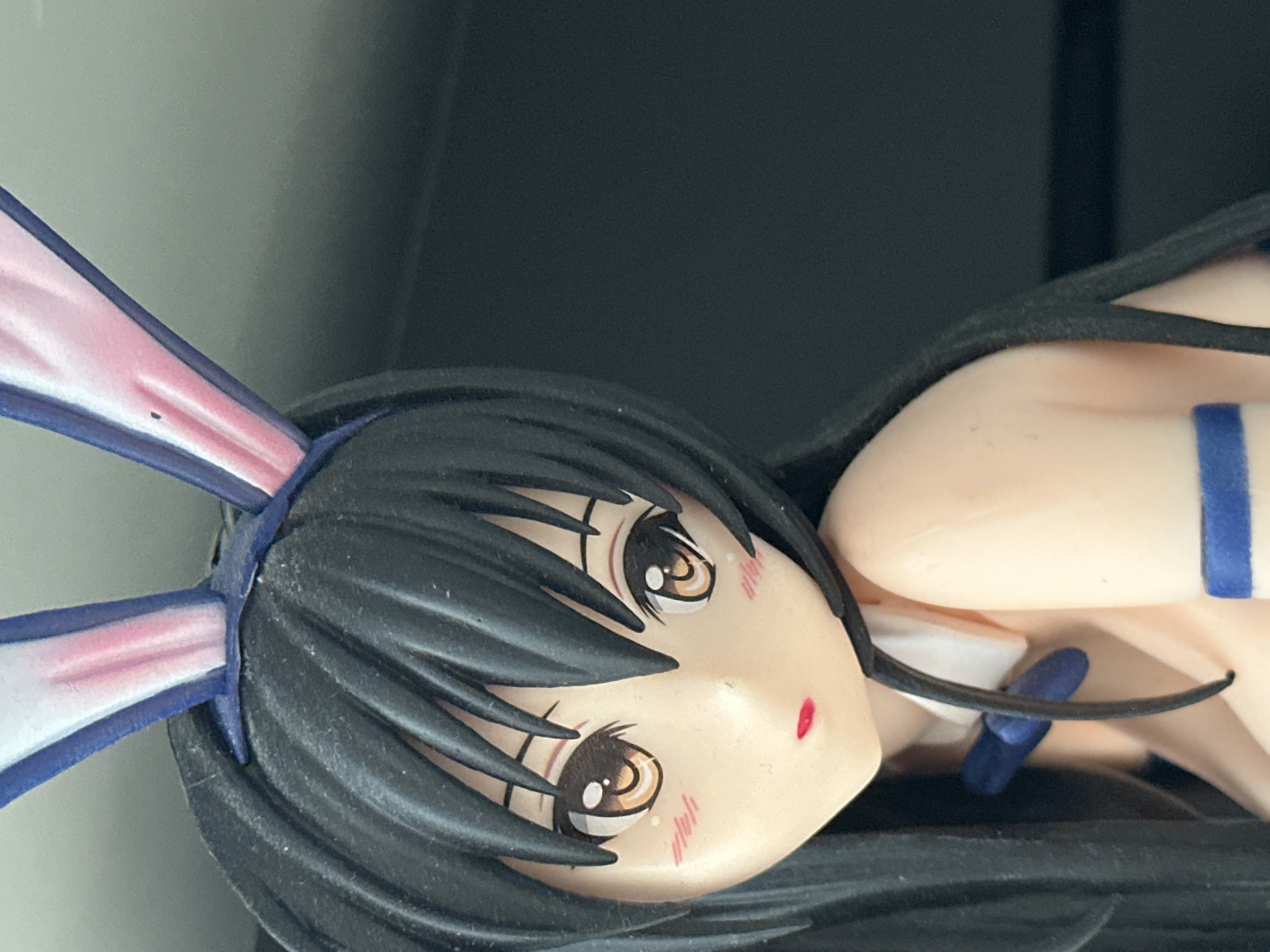 This is a close-up, high-angle, eye-level shot of an anime-style figurine. The figurine is a young woman with long black hair, large brown eyes with white pupils, rosy cheeks, and red lips. She is wearing a peach-colored outfit with a white collar and a blue bow tie. She also has pink and blue bunny ears. The background is dark and out of focus, with a lighter gray area on the left side. The lighting is bright, highlighting the details of the figurine. There is no visible connection to Nevinnomyssk, Russia, or any urban environment.