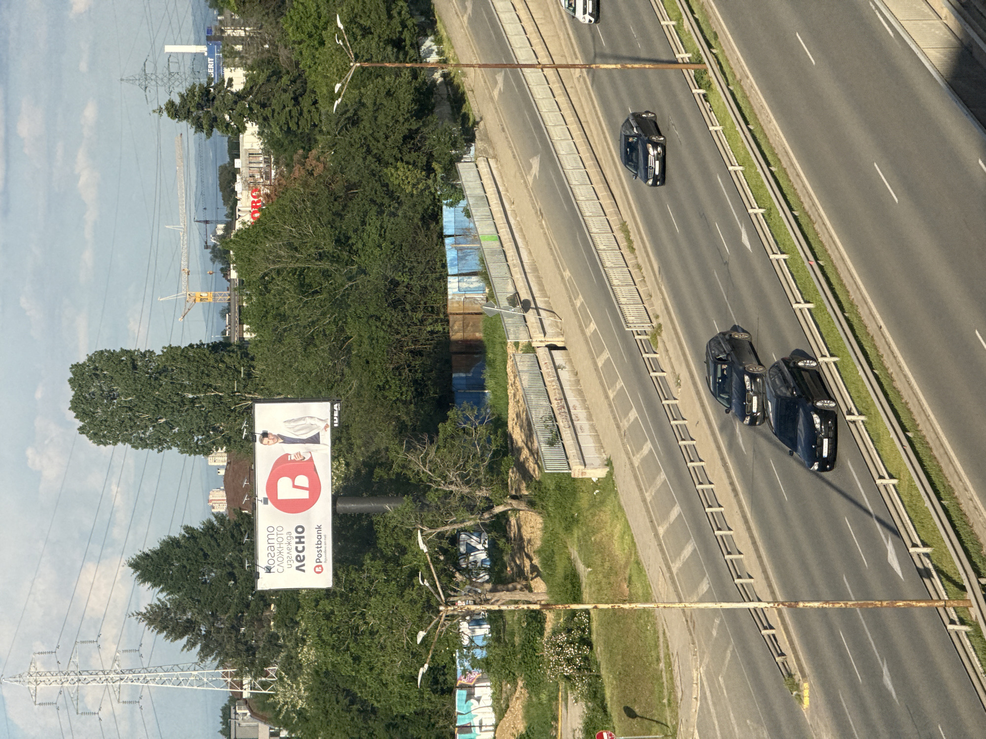 The image depicts a section of a multi-lane highway in Sofia, Bulgaria. The road, likely a major thoroughfare, features multiple lanes separated by concrete barriers and green vegetation. Cars are visible traveling on the road. Adjacent to the highway is a patch of greenery with trees, power lines, and a large billboard advertisement for Postbank. In the distance, more buildings and structures are visible, including a crane.