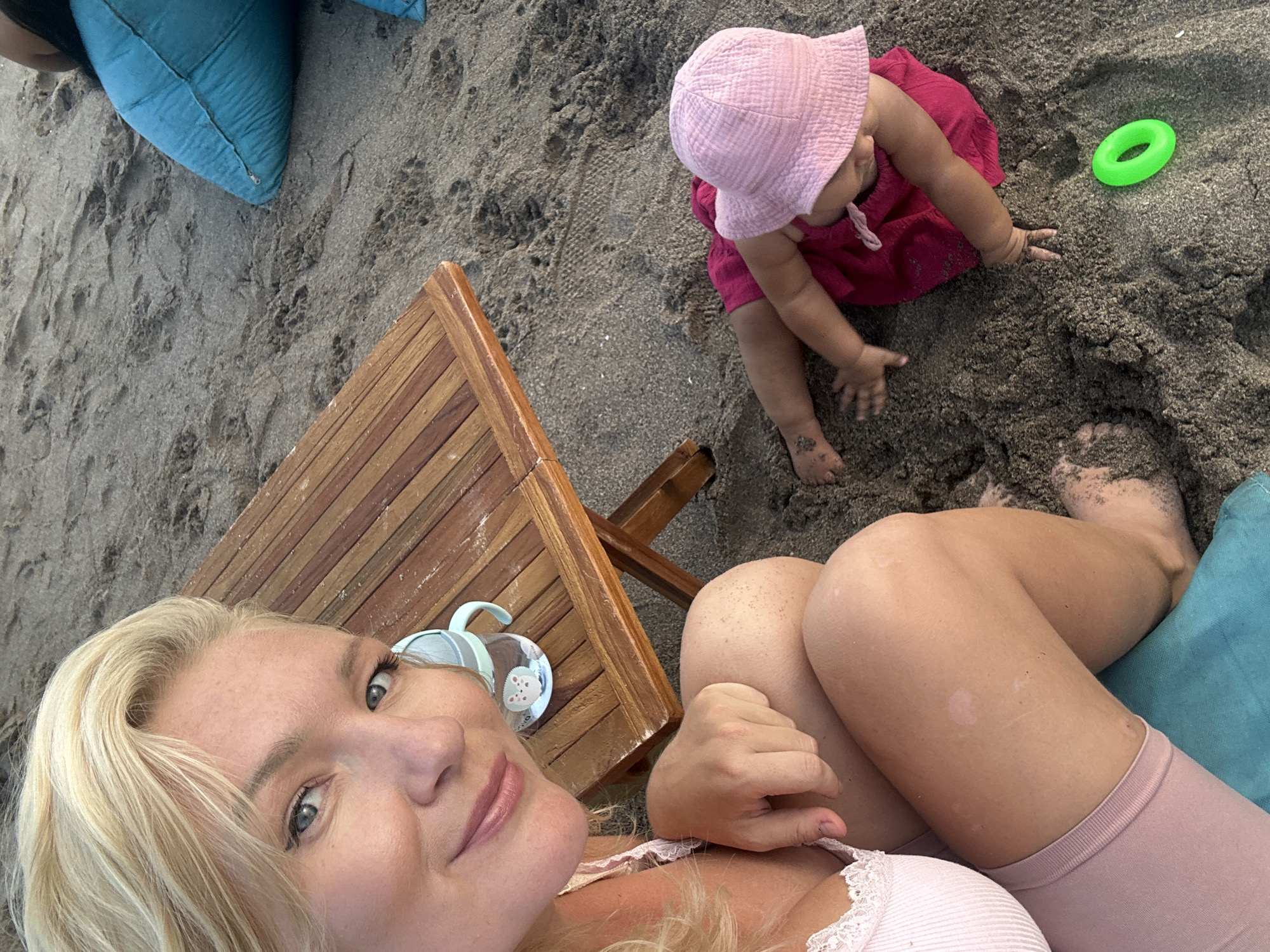 The image shows a woman and baby on a beach in North Kuta, Indonesia. The woman is fair-skinned, with blonde hair, and is lying on a turquoise pillow next to a small wooden table. She is wearing a pink top and pink shorts. A baby in a pink hat and red dress is playing in the sand near a green toy ring. The sand is dark grey. The baby has some sand on their feet. A blue water bottle is on the table next to the woman.