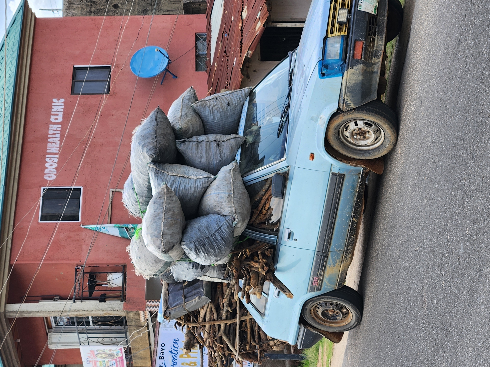 A blue car heavily loaded with large bags and wood is parked on the side of a road. The car's front tires are visible. Behind the car is a red building with the words "ODOSI HEALTH CLINIC" written on it, and a blue satellite dish is mounted on the building's exterior. The street is labeled as Rukuba Road in Nigeria.