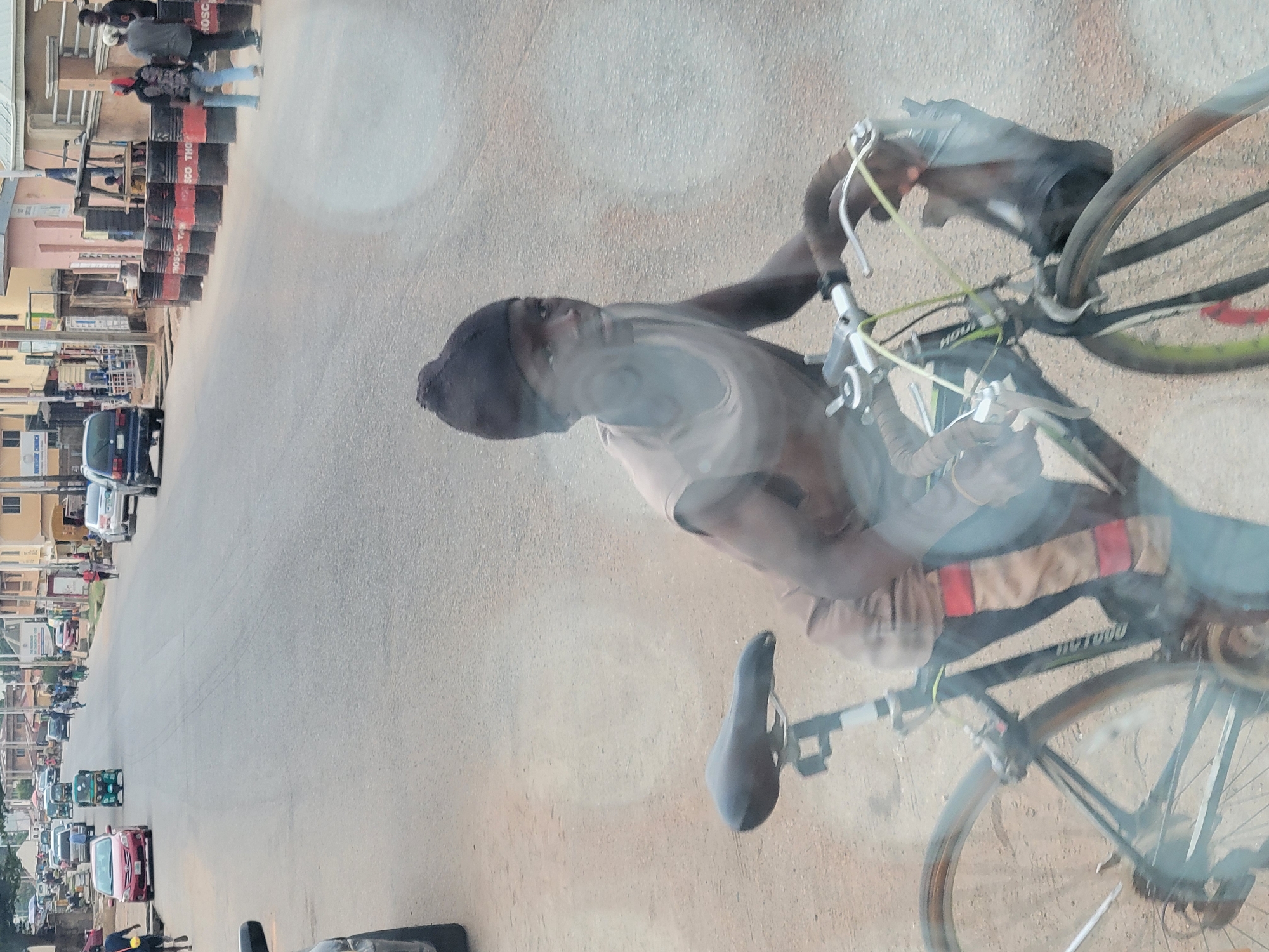 A photograph of a street in Jos, Nigeria, depicts a man on a bicycle riding along the right side of the road. He is wearing a dark cap and a light-colored shirt, carrying a bag on the handlebars. The road is lined with buildings and shops on the left, with a line of black barrels acting as a makeshift barrier. Traffic, including cars and tuk-tuks, moves in the opposite direction. Pedestrians are visible on the sidewalk. The photograph is taken from an elevated position, likely from within a vehicle.