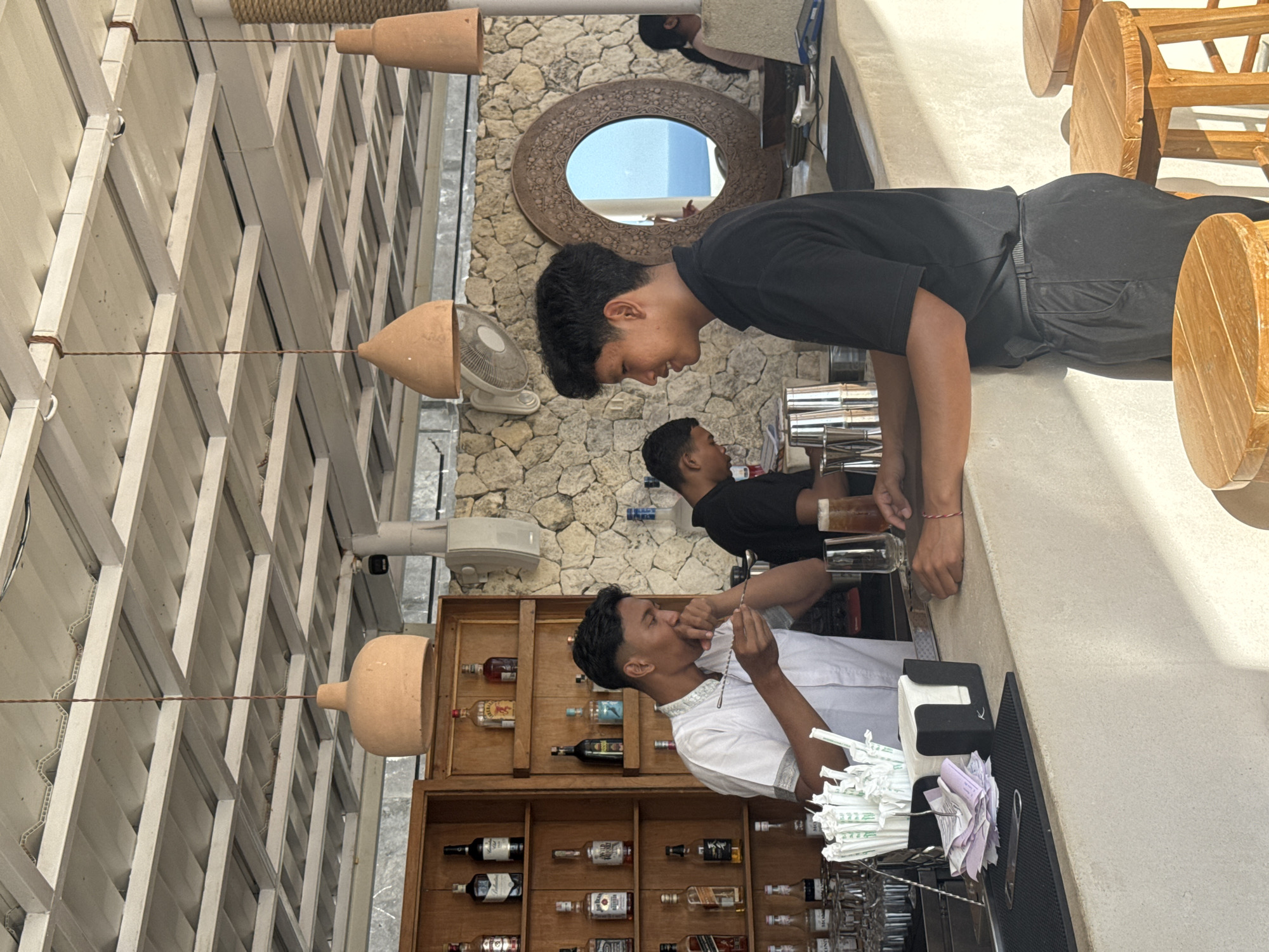 Here is a description of the image:
The image depicts an outdoor bar setting in Kuta Selatan, Indonesia. Three male bartenders are visible. The first bartender is wearing a white shirt and is tasting a drink with a spoon. Another bartender is wearing a black shirt, and a third is pouring a beverage. The bar is stocked with a variety of liquor bottles and glassware. Above the bar are hanging terracotta pots and a white pergola. A round mirror is mounted on the stone wall. Wooden chairs are visible on the right side of the frame.