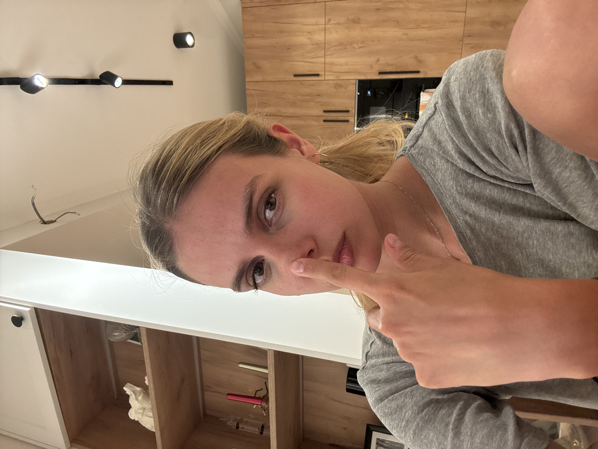 A woman is indoors with her finger touching her nose. She wears a gray shirt and a gold necklace. She has blonde hair pulled back in a ponytail. Behind her is a kitchen with light wood cabinets and a white countertop. A shelf with various items is also visible. Overhead, there is track lighting on a white ceiling. The location is Unknown, Unknown.