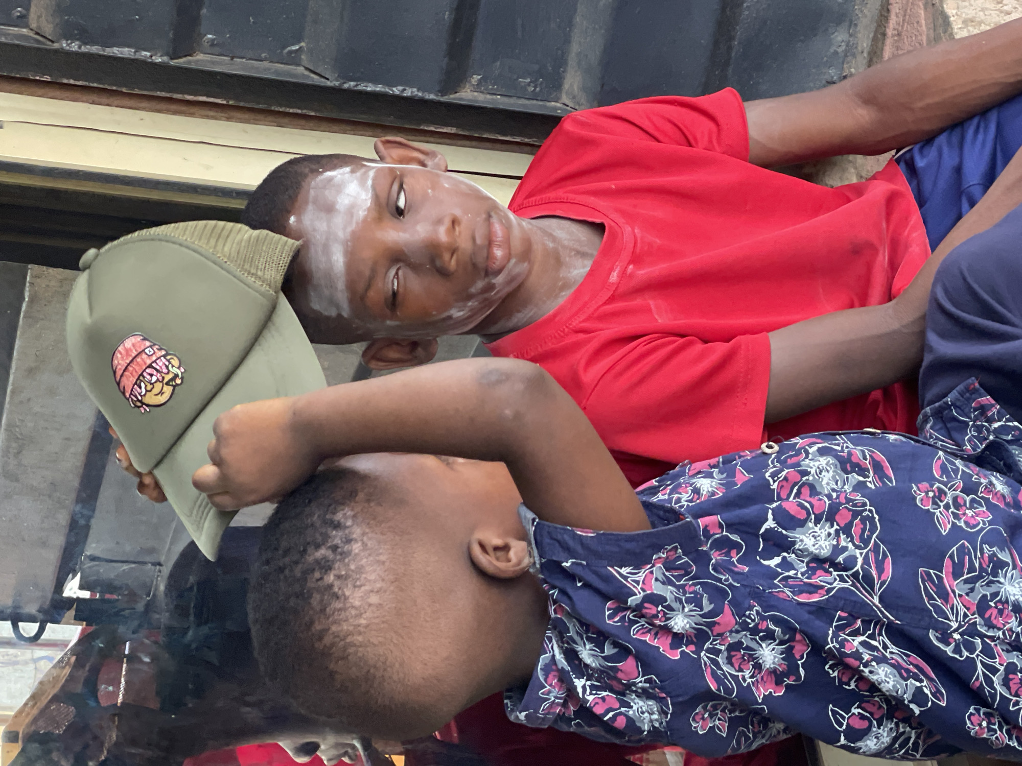 Two young African boys are lying down. One boy is wearing a red t-shirt and blue shorts, with white powder smeared on his face. The other boy is wearing a dark blue shirt with a floral pattern and is holding a green baseball cap with a cartoon patch. The scene appears to be outdoors in Onitsha, Nigeria, given the setting. The image is angled and may have been reflected off a surface.