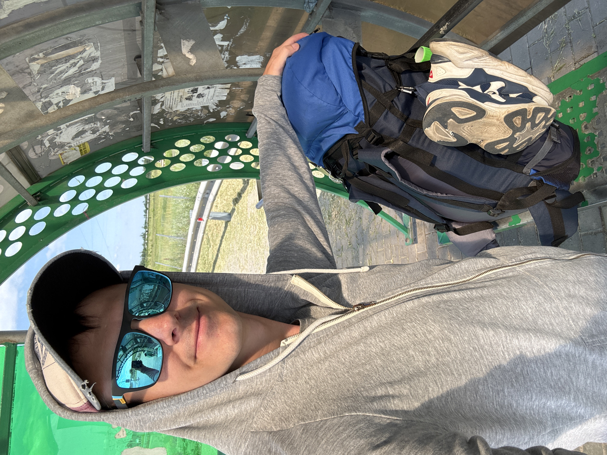 A man wearing a grey hooded sweatshirt, a baseball cap, and blue mirrored sunglasses stands outdoors next to a blue backpack attached to a green metal structure with circular cutouts. His right arm rests on the backpack. In the reflection of his sunglasses, a hand holding a phone and the man's face are visible. The backpack has a pair of dirty white and blue athletic shoes attached to its side. The background shows a grassy field and a blue sky. The structure appears to be a bus stop or shelter, with torn posters on its interior wall. The location is identified as МТФ №2 колхоз 40 лет Октября, Russia.