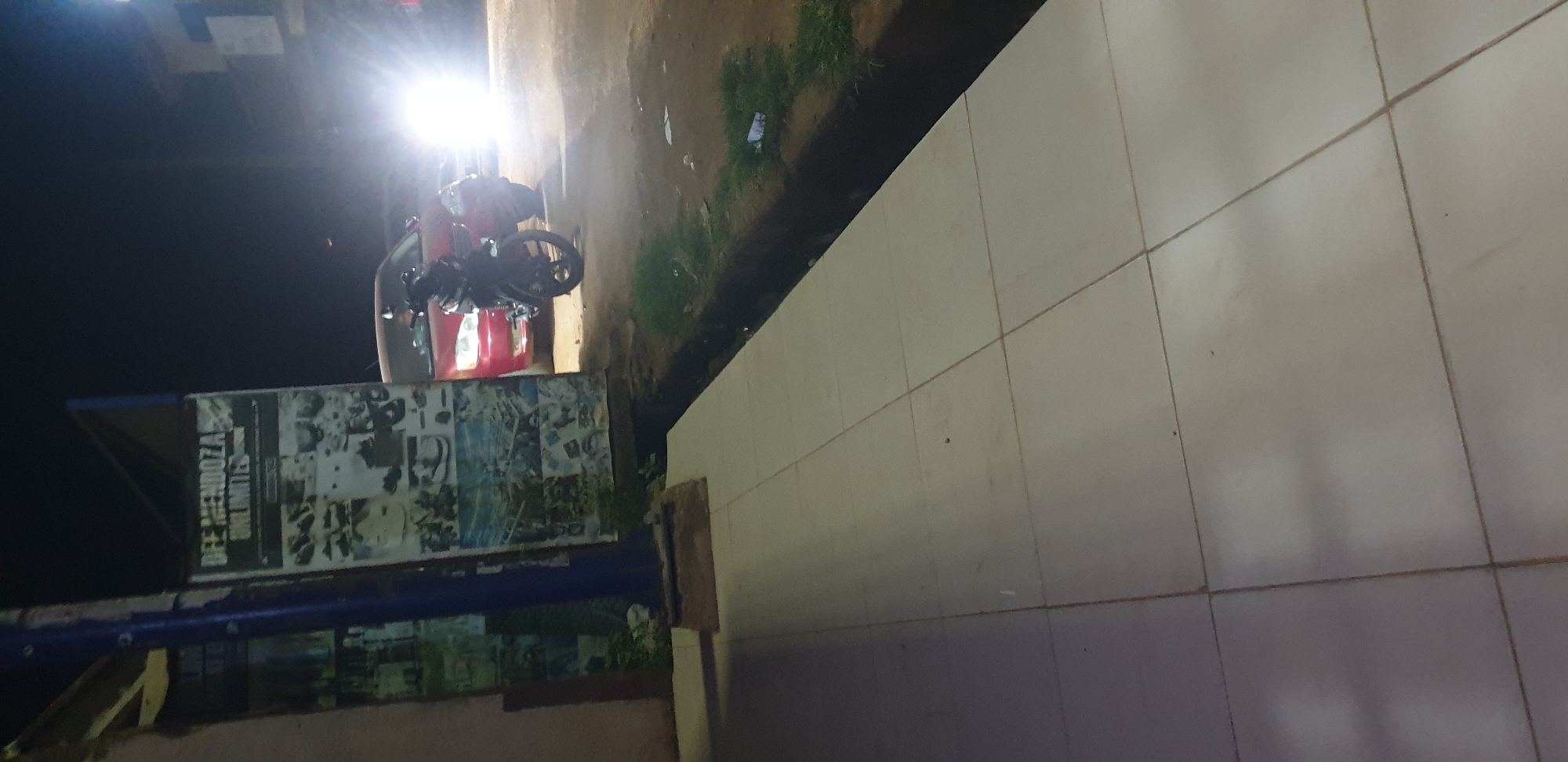 The image captures a nighttime street scene in Narayi, Nigeria. In the foreground, a light-colored tiled sidewalk extends from the lower-left to the middle-right of the frame, showing faint discolorations and grid lines. A dark, narrow drainage ditch separates the sidewalk from a strip of sparse grass and dirt. In the midground, a blue vertical pole stands adjacent to a wall adorned with multiple posters; one on the right is legible, displaying "DEE MENDOZA UNLIMITED" at its top, above several images of footwear. To the right of these structures, a red sedan is partially visible, with a dark-colored motorcycle parked or stationary directly in front of it, facing right. Further right in the midground and background, bright vehicle headlights emit a strong glare, obscuring details of additional vehicles and distant background structures. No individuals are discernible within the frame.