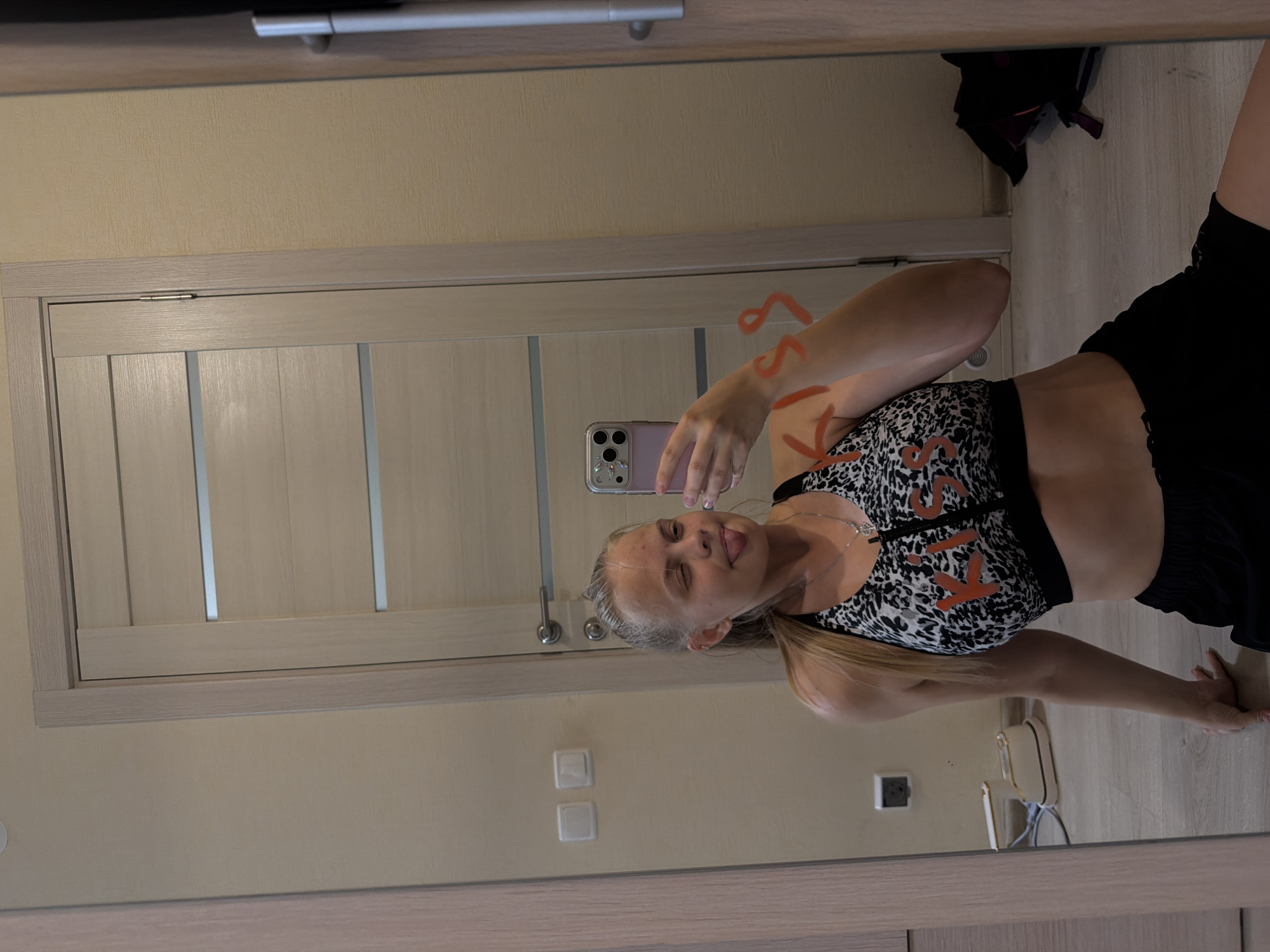 A young woman with blonde hair tied in a ponytail is taking a selfie in a mirror. She is wearing a black and white leopard print sports bra and black shorts. She is sticking her tongue out and winking. The word "KISS" is written in red on the mirror. She is sitting on the floor in front of a light-colored door. There are light switches on the wall to the left of the door. In the reflection, a white and gold appliance is visible on the floor. To the right of the door, a dark bag is on the floor. The room appears to be a residential interior in Izhevsk, Russia.