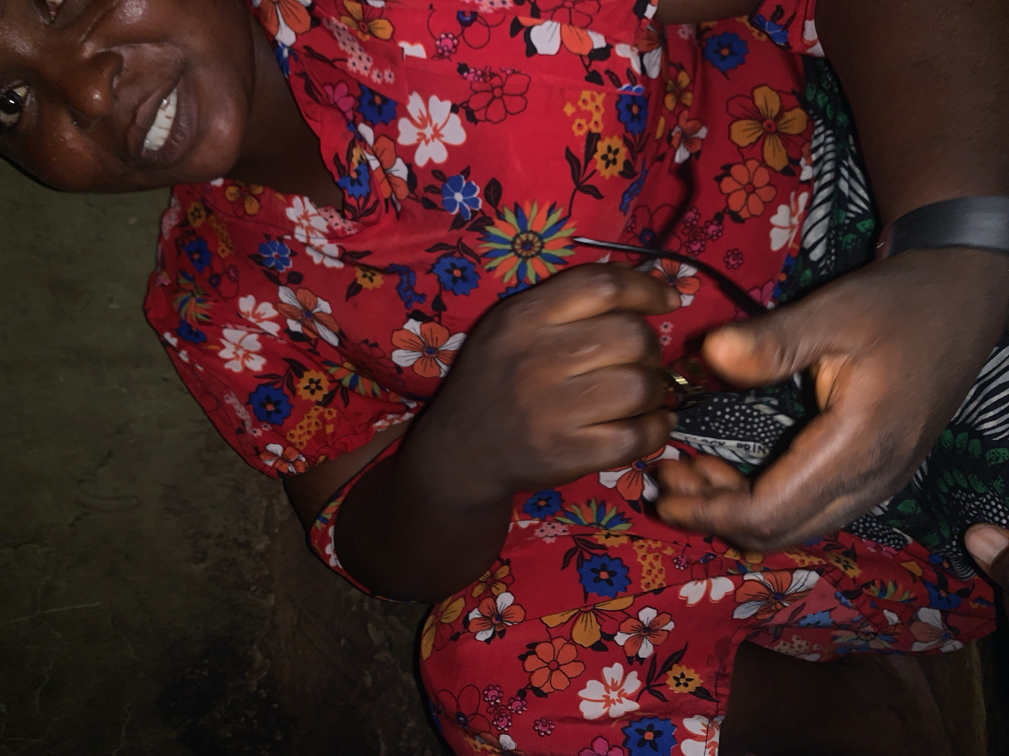 In Project, Nigeria, an individual wearing a red floral-patterned dress and a black wristband is seated against a dark, textured wall. Their hands are clasped in front of them, holding a dark object that appears to be a pair of glasses. The individual's face is partially visible, showing dark skin, a wide smile, and visible teeth. The lighting is dim, with a spotlight effect on the person's face and hands, suggesting an indoor setting or nighttime environment.