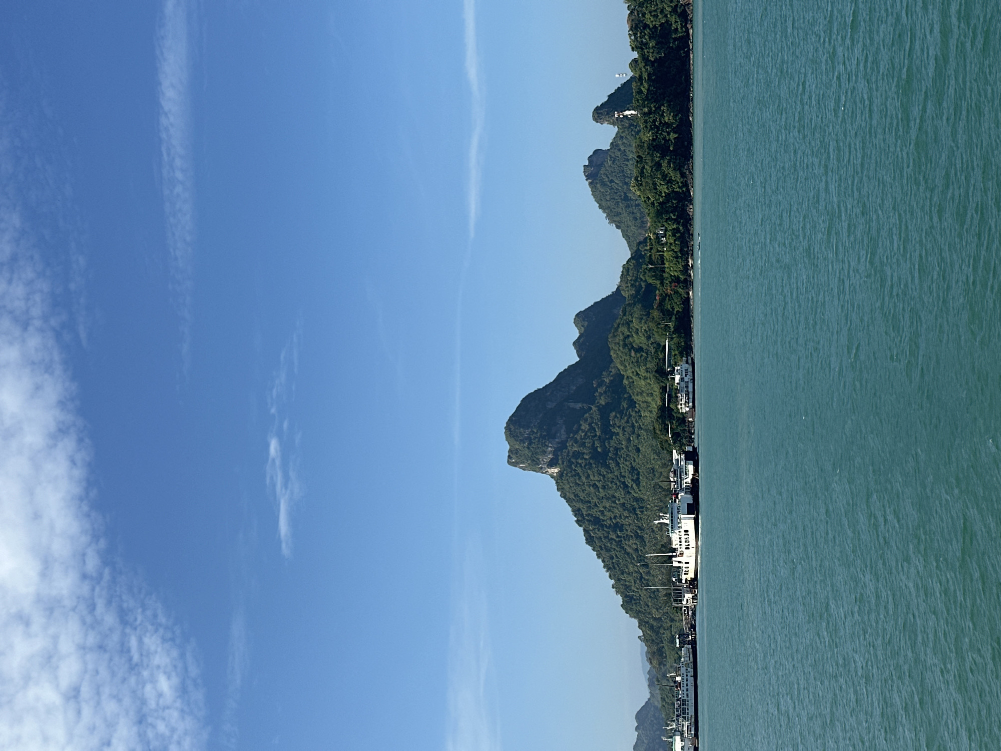 The image shows a view of the city of Don Sak, Thailand, with a body of water in the foreground and lush green hills in the background. Several ferries are docked along the coast, suggesting this is a port or ferry terminal. The water is a vibrant turquoise, and the sky is a clear blue with a few wispy clouds. The hills are densely covered in trees, with some rocky outcrops visible. The overall scene depicts a scenic coastal landscape with active maritime traffic.