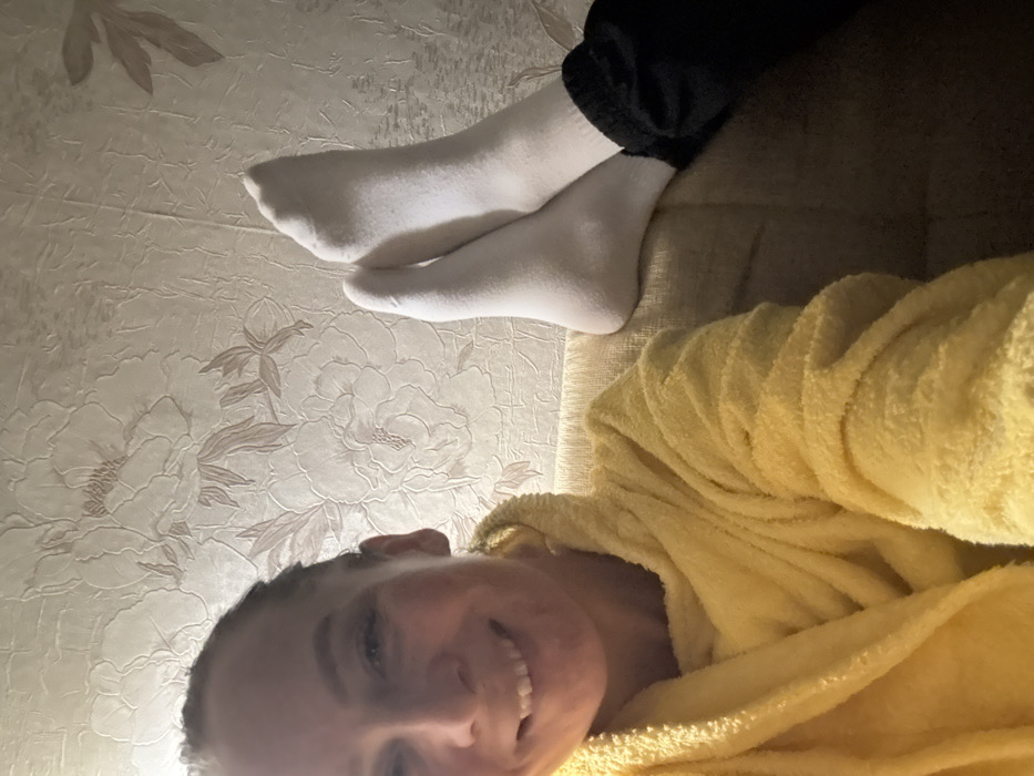 The image depicts an indoor scene in Pavlovsky, Russia. An adult female, with her hair pulled back and smiling, occupies the left side of the frame. She wears a yellow, textured bathrobe. Her feet, clad in white ankle socks and dark trousers, are elevated and rest on a light brown fabric sofa in the right portion of the image. The background consists of light-colored wallpaper featuring a textured floral pattern. The scene is illuminated by artificial light.