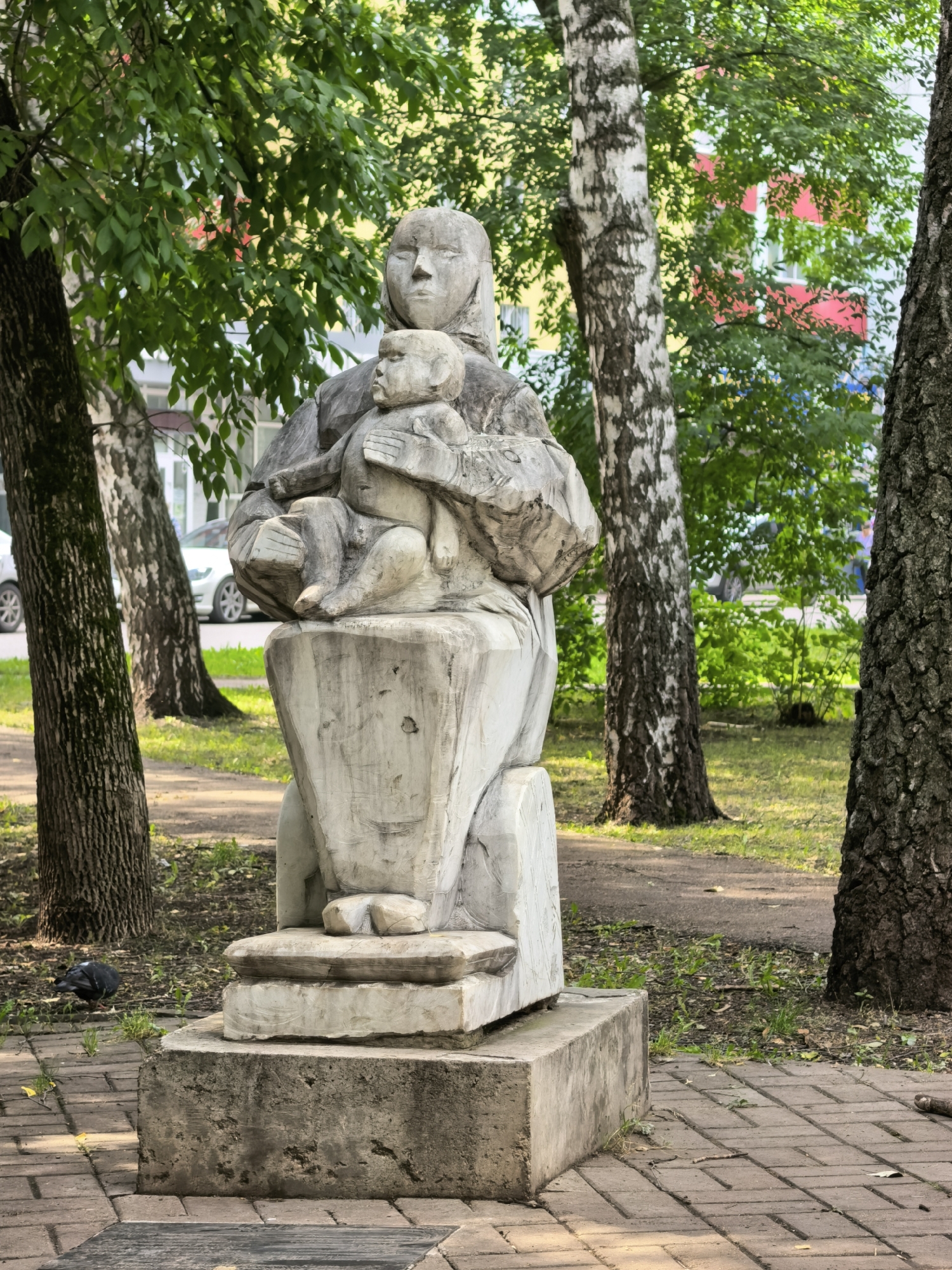 An outdoor, eye-level, full-shot photograph depicts a weathered, light-gray stone sculpture of a mother holding a child. The sculpture is situated on a multi-tiered stone pedestal in what appears to be a park or public square. The mother is depicted with a serene, downward gaze, her head covered by a headscarf. The child is cradled in her arms, its face turned towards the viewer. The sculpture exhibits a rough, hand-carved texture, with visible chisel marks.
Behind the sculpture, a paved walkway made of reddish-brown brick pavers is visible. A single pigeon is present on the pavers to the left of the sculpture. Lush green trees with thick trunks surround the sculpture, with their leaves forming a dense canopy overhead. In the background, beyond the trees, a building with yellow and red facades is visible, suggesting an urban environment. The ground around the sculpture is covered with grass and scattered leaves. The overall lighting suggests a bright, possibly sunny day. This sculpture is located on the territory of the Republic of Bashkortostan, in the city of Sterlitamak, Russia.