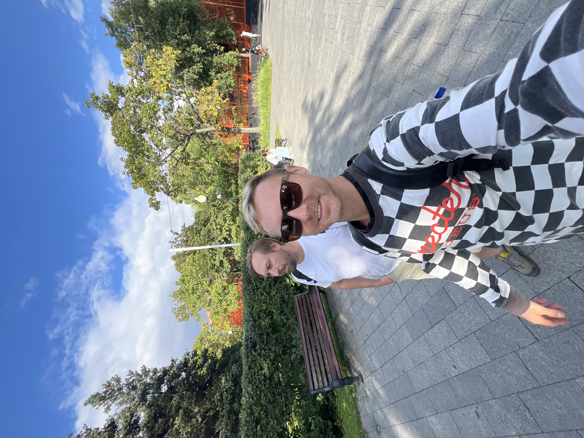 Two men are taking a selfie in a park. The man in the foreground is wearing a black and white checkered long-sleeved shirt and sunglasses. He is smiling at the camera. The man behind him is wearing a white t-shirt and khaki shorts, and he is looking to the side. The background shows a paved walkway, a wooden bench, a hedge, trees, and a bright blue sky with scattered clouds. In the distance, there are a few other people, including one on a bicycle, and a red fence. The scene appears to be in a park in Moscow, Russia, possibly Gorky Park or a similar recreational area given the sunny weather, paved pathways, benches, and greenery.