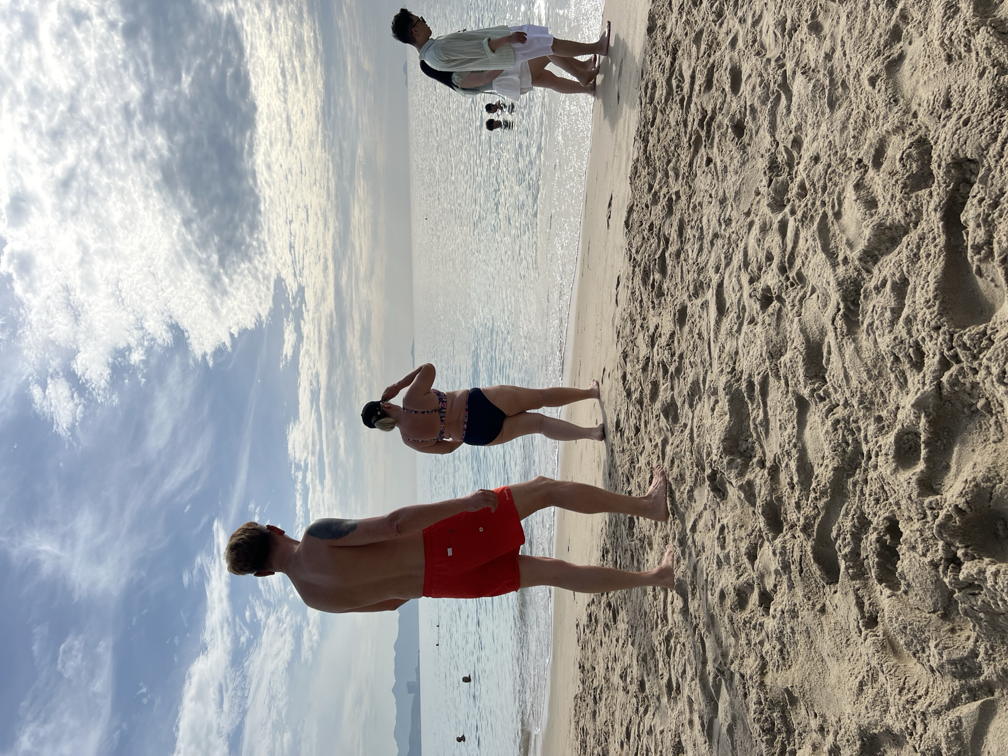 This is a medium-shot photograph taken on a sandy beach, likely in Nha Trang, Vietnam, given the presence of distant hills and urban structures indicative of the city's coastline. The sky is partly cloudy with wispy white clouds against a blue backdrop. The sand in the foreground is textured with footprints and ripples.
In the mid-ground, several people are present. On the left, a shirtless man with blonde hair stands facing the ocean, wearing red swim trunks. To his right, a woman in a dark bikini with a patterned top is also facing the water, adjusting her hat. Further to the right, a group of people walk along the shoreline. A man in a light green shirt and white shorts is walking with a woman in a white skirt and a light-colored top. Behind them, two children are partially submerged in the shallow water, appearing to play. In the far distance, the calm ocean stretches to the horizon, meeting a hazy sky. The water is light blue with gentle ripples. The overall scene depicts a relaxed day at the beach with people enjoying the coastal environment.