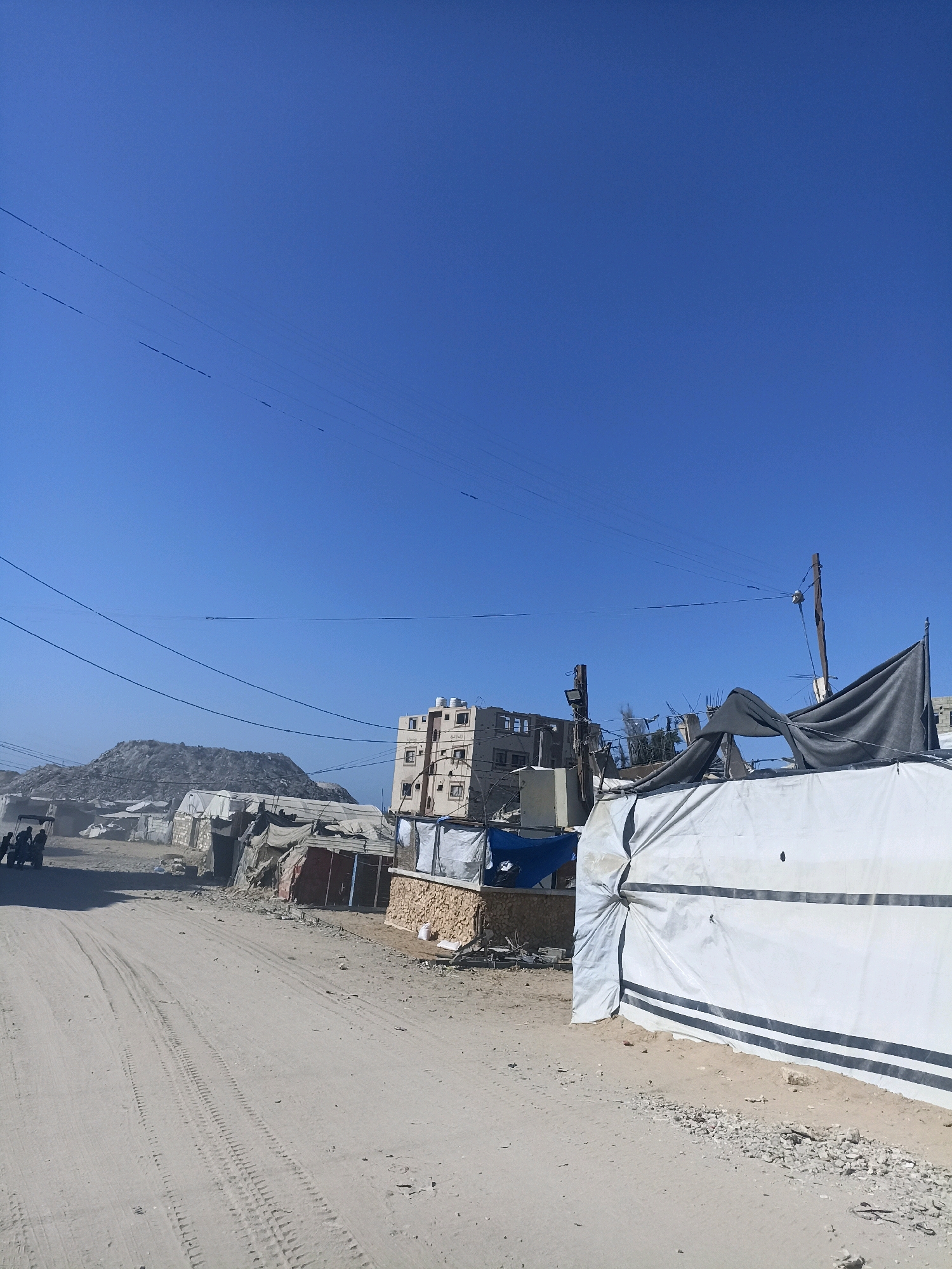 This image depicts a dusty, outdoor scene in the Gaza Strip, Palestinian Territories, characterized by makeshift structures, debris, and a clear blue sky with overhead power lines. In the foreground, a dirt road shows tire tracks, suggesting recent vehicle activity. To the left, a few people are visible near a vehicle, possibly a golf cart or utility vehicle.
The middle ground is a jumble of temporary dwellings constructed from tarps, corrugated metal, and what appear to be cinder blocks. Some structures are covered with white tarps, while others are made of darker fabrics. A large pile of rubble or debris forms a hill in the background to the left.
A multi-story building, appearing damaged with visible damage to its facade and windows, stands towards the center-right of the image. In front of this building and extending to the right, more temporary structures are visible, including a large white tarp structure with dark stripes. The environment is arid and sandy, with scattered rocks and debris on the ground. The overall impression is one of a settlement or displacement camp in a damaged urban or semi-urban area.