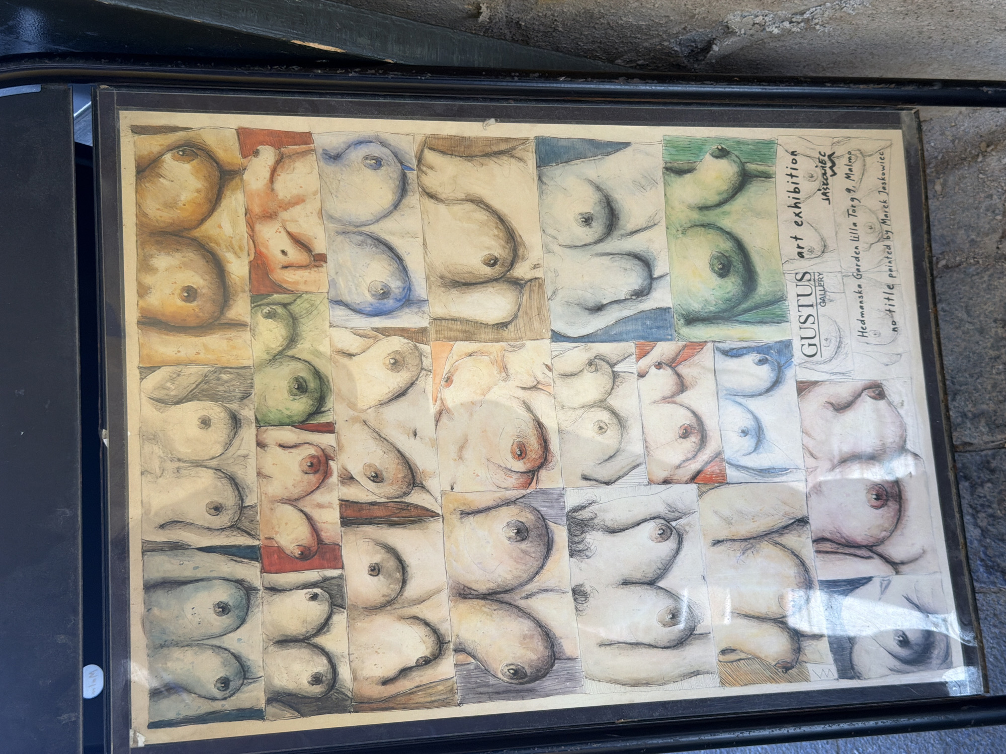The image shows a framed exhibition poster featuring a collage of watercolor and pencil drawings of female breasts. The poster is displayed outdoors, leaning against a textured stone wall, suggesting it might be in a public space. The poster is titled "GUSTUS GALLERY art exhibition" and includes text indicating the location as "Hedmanska Garden Lilla Torg 9, Malmö" and "no title painted by Marek Jaskowiec". Lilla Torg is a well-known historic square in Malmö, Sweden, famous for its cobblestone streets and well-preserved buildings. The artwork itself is a grid of numerous individual drawings of breasts, rendered in varying colors and styles. Some drawings are colored with watercolors, while others are sketched with pencil. The overall presentation is that of an art display.