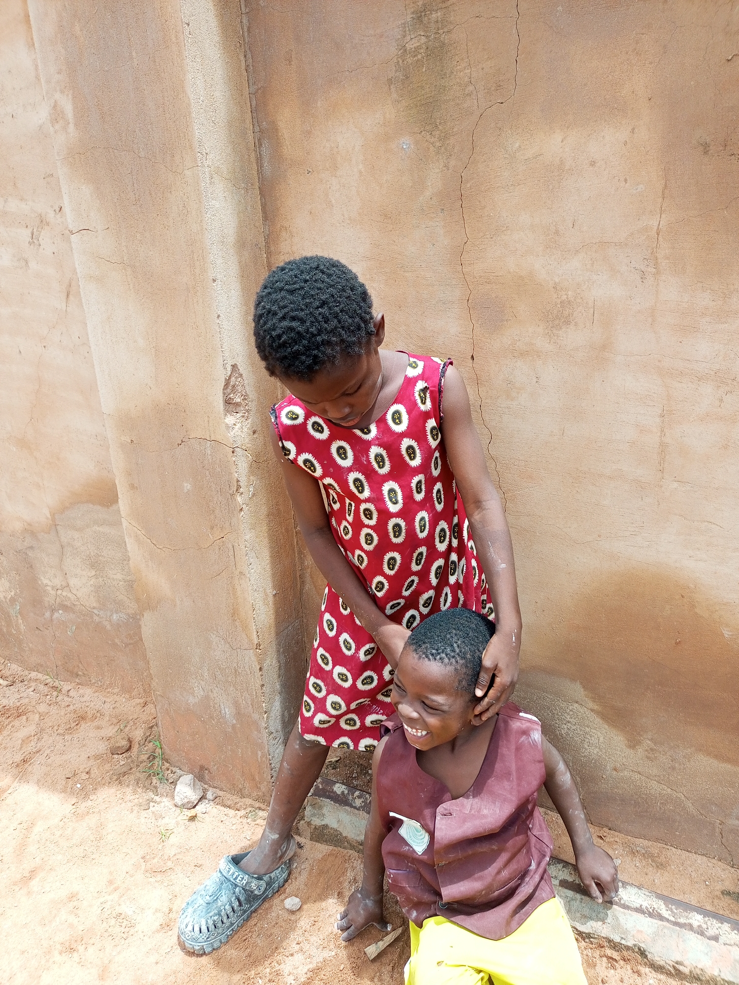 Two young children are in the city of Tempure, Nigeria. The child on the left, wearing a red dress with a white floral pattern, stands and appears to be gently touching the head of the child on the right. The child on the right, wearing a maroon vest over a yellow garment, is sitting and laughing. They are positioned in front of a weathered, textured wall. The ground is sandy and appears to be outdoors.