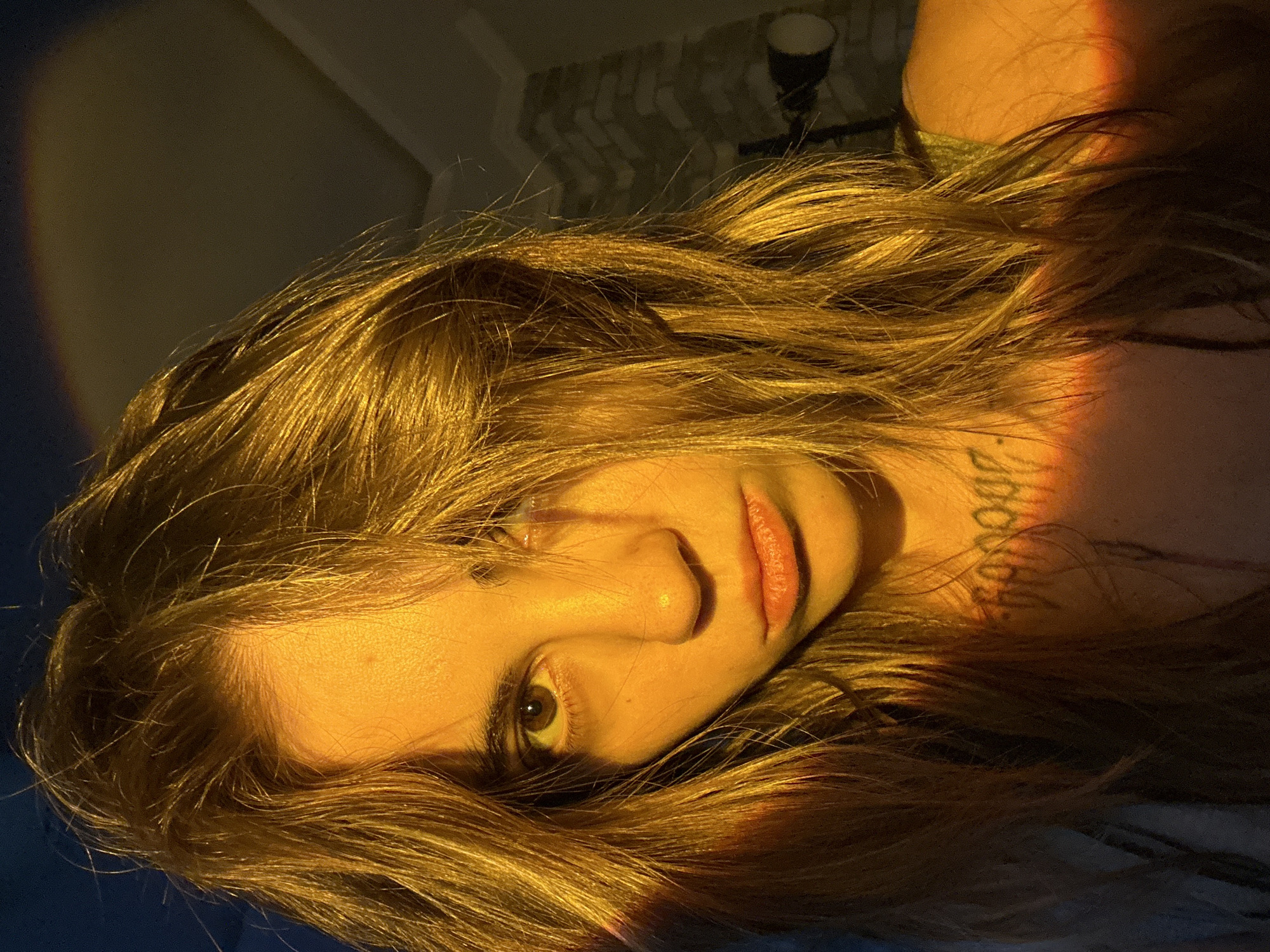 The image is a close-up, low-angle, self-portrait of a young woman with long, light brown hair. She is illuminated by a warm, golden light that casts shadows across her face, highlighting her brown eyes, nose, and lips. A subtle, reddish-orange light also streaks across the left side of her face and hair, suggesting a colored light source. She has a neutral expression. A tattoo is visible on her neck. The background appears to be an indoor environment with a brick wall and a light fixture visible on the right. There is no information in the image to suggest a location in Moscow, Russia.