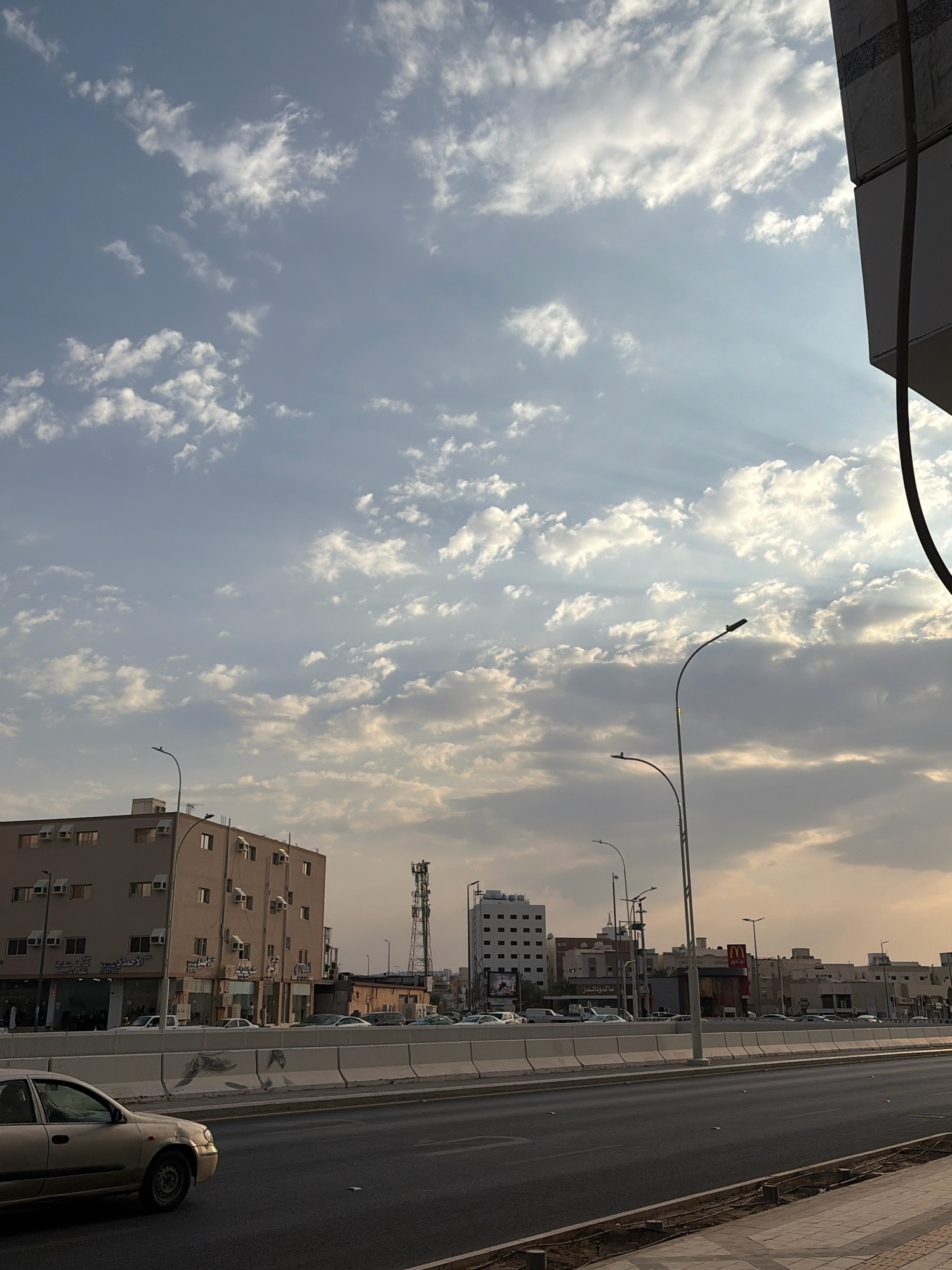 The image depicts a street scene in Medina, Saudi Arabia, during daylight. The sky is filled with scattered clouds, with sunbeams radiating through them. In the foreground, a multi-lane road is visible, with a light-colored car in the left lane and a concrete barrier separating the road from a sidewalk. Along the right side of the road, a sidewalk is present. Across the road, there are buildings, including a multi-story building with many windows and air conditioning units, and further down, a McDonald's restaurant with its prominent signage. Communication towers are also visible in the background. The overall impression is of a typical urban environment with moderate traffic and commercial activity. There are no people clearly visible in the image, and the focus is on the cityscape and the sky.