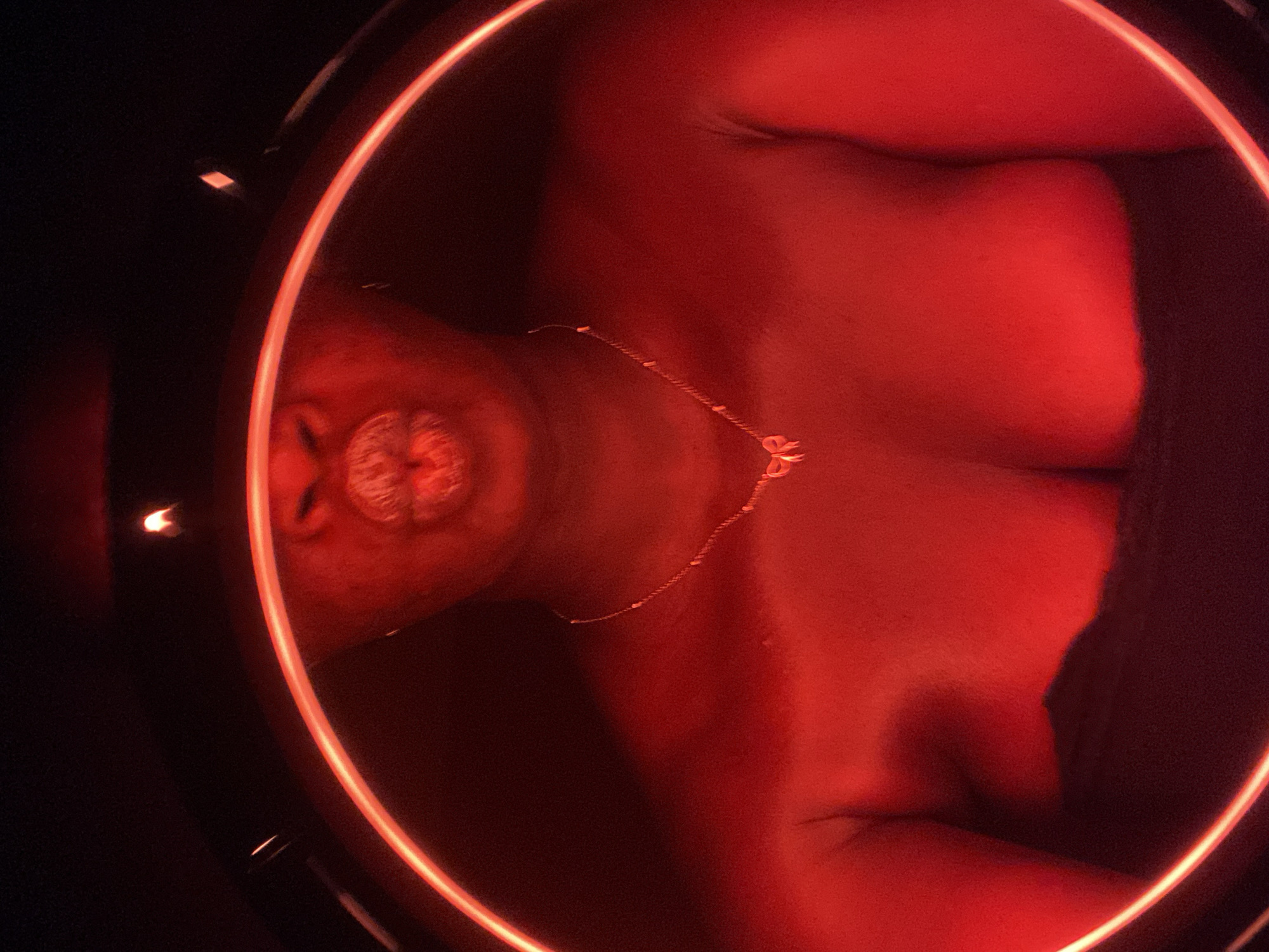 A close-up image depicts a person lying down within a circular, illuminated red ring. The individual is fair-skinned and wears a delicate gold necklace. Facial features, including the nose and mouth, are prominent. The person's upper chest and shoulders are visible. The background is dark and indistinct. The location is unknown.