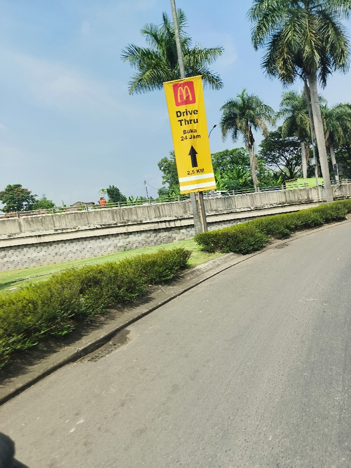 The image shows a signpost located on the side of a road in Padalarang, Indonesia. The signpost is attached to a metal pole and has a yellow background with a red McDonald's "M" logo in the top left corner. The text on the sign reads "Drive Thru Buka 24 Jam" (Drive Thru Open 24 Hours) and "2.5 KM" below an upward pointing arrow. There are bushes in the foreground and a gray concrete wall behind the signpost. The road appears to curve to the right. There are trees in the background. The coordinates provided are consistent with a location in Padalarang, Indonesia. The image was taken from a moving vehicle looking to the right. The weather appears to be clear.
