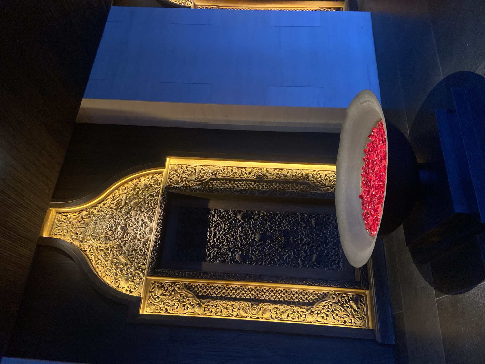 The image shows an ornate wall decoration inside a building in Kuta Selatan, Indonesia. The wall features a large, elaborately carved wooden frame with intricate gold-colored detailing. Inside the frame is a panel with a dark background and a filigree design. To the right of the frame is a tall, grey stone vessel filled with red flower petals. The vessel sits on a dark-colored pedestal. Above the wall decoration is a section of a blue wall. The floor appears to be dark-colored stone or tile. There is no indication of people in the image.