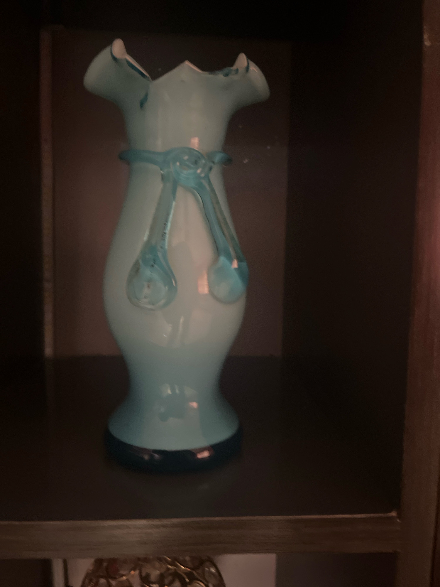 A light blue glass vase with a ruffled rim and a dark blue base is displayed on a dark brown shelf. The vase has a decorative blue glass ribbon tied around its neck, with two teardrop-shaped elements hanging from it. The background is dark and out of focus, suggesting an indoor setting, possibly a shelf in a room. The image does not contain any information that would allow for its location to be identified within Jos, Nigeria, or any specific city.