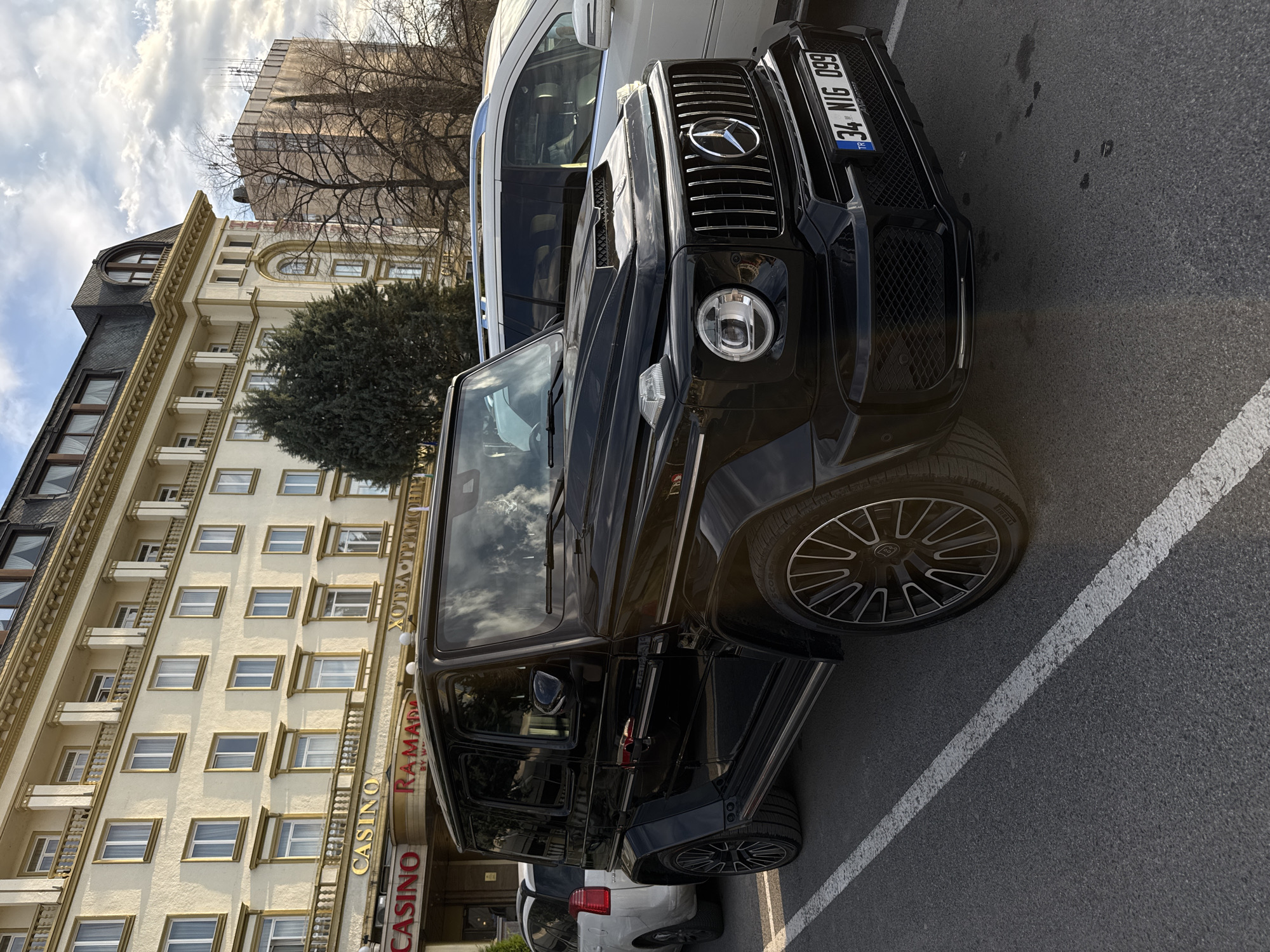 The image shows a black Mercedes-Benz G-Class parked on a street in Plovdiv, Bulgaria, in front of the Ramada by Wyndham Plovdiv Trimontium Casino. The car is parked parallel to the curb, next to a white van. The building behind the car is a tall, beige-colored building with numerous windows and balconies. The sky is partly cloudy. The license plate of the G-Class is visible, reading "34 NIG 099". The casino signage is partially visible.