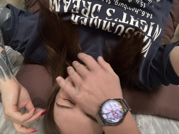 The image displays two individuals in an indoor setting, likely a residential space, viewed from above. One individual's left hand, wearing a dark metallic smartwatch with a circular, illuminated digital face displaying red indicators, is placed over the lower facial area of a second individual. The fingers cover the nose and mouth region, while the thumb rests on the cheek.
The second individual, appearing to be female, is positioned supine, with long, dark brown hair spread out. She wears a dark blue short-sleeved t-shirt with white text partially legible as "SUMMER NIGHT'S DREAM 14.6., Hamburg, St". Her right eye is visible, appearing partially closed or squinted, and her left forearm displays extensive dark-ink tattoos, potentially depicting floral or abstract designs. Her left hand's fingernails are painted red.
The subjects are situated on a surface consistent with a bed or sofa, featuring a brown pillow under the second individual's head and a light-colored, patterned fabric visible in the background. The scene is located in Chornomorskoe, Ukraine.