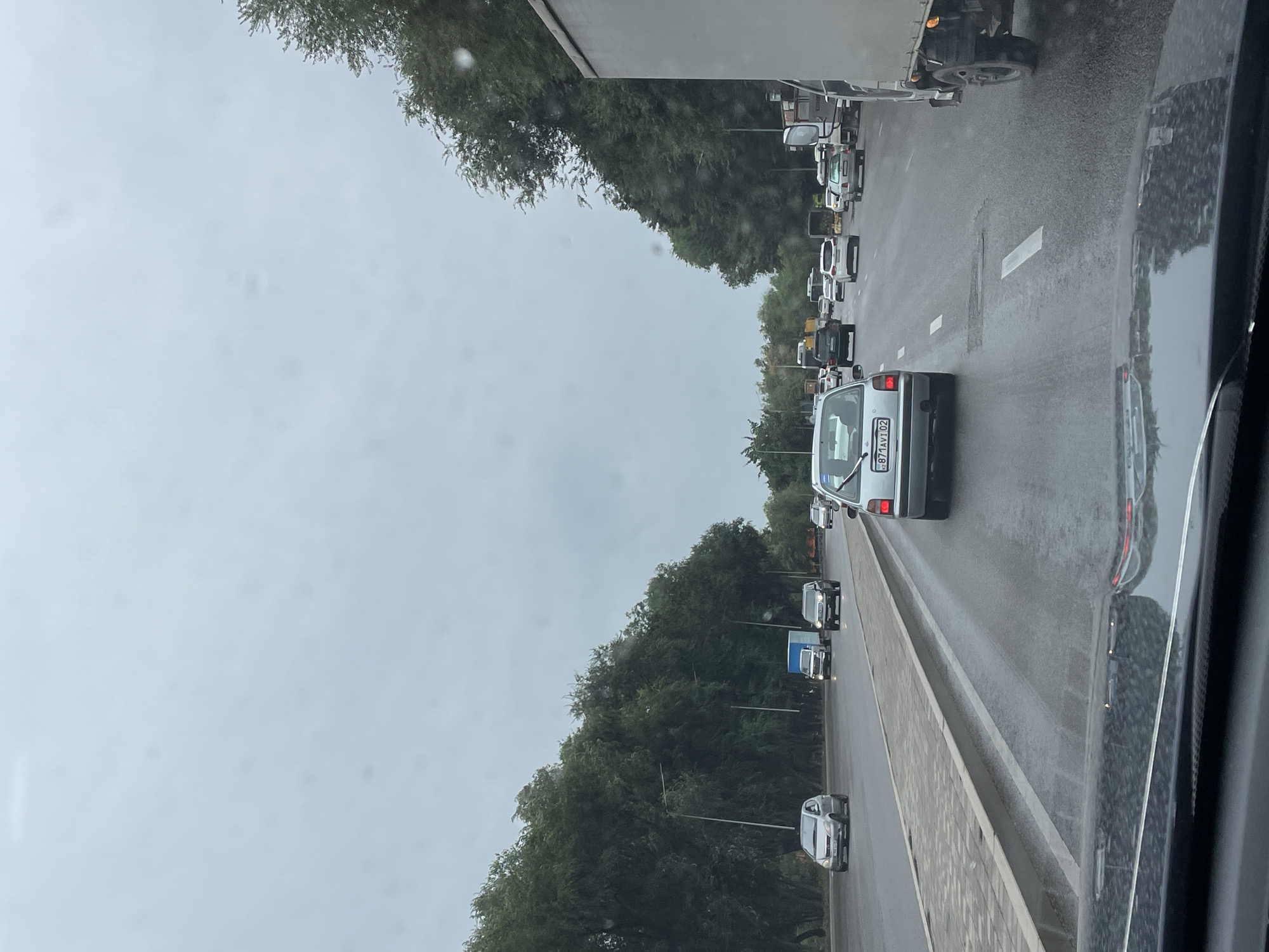 The image depicts a street scene in Almaty, Kazakhstan, on a cloudy day. A multi-lane road is filled with cars, indicating traffic. On the left side of the road, a paved walkway is visible, bordered by a curb and then a dense line of green trees. Streetlights are interspersed among the trees. On the right side, another line of trees forms a boundary. The road surface appears wet, suggesting recent rain. Several cars are moving in the same direction, with a larger truck visible in the right lane. The foreground shows the dashboard and windshield of a vehicle, with reflections on the windshield.